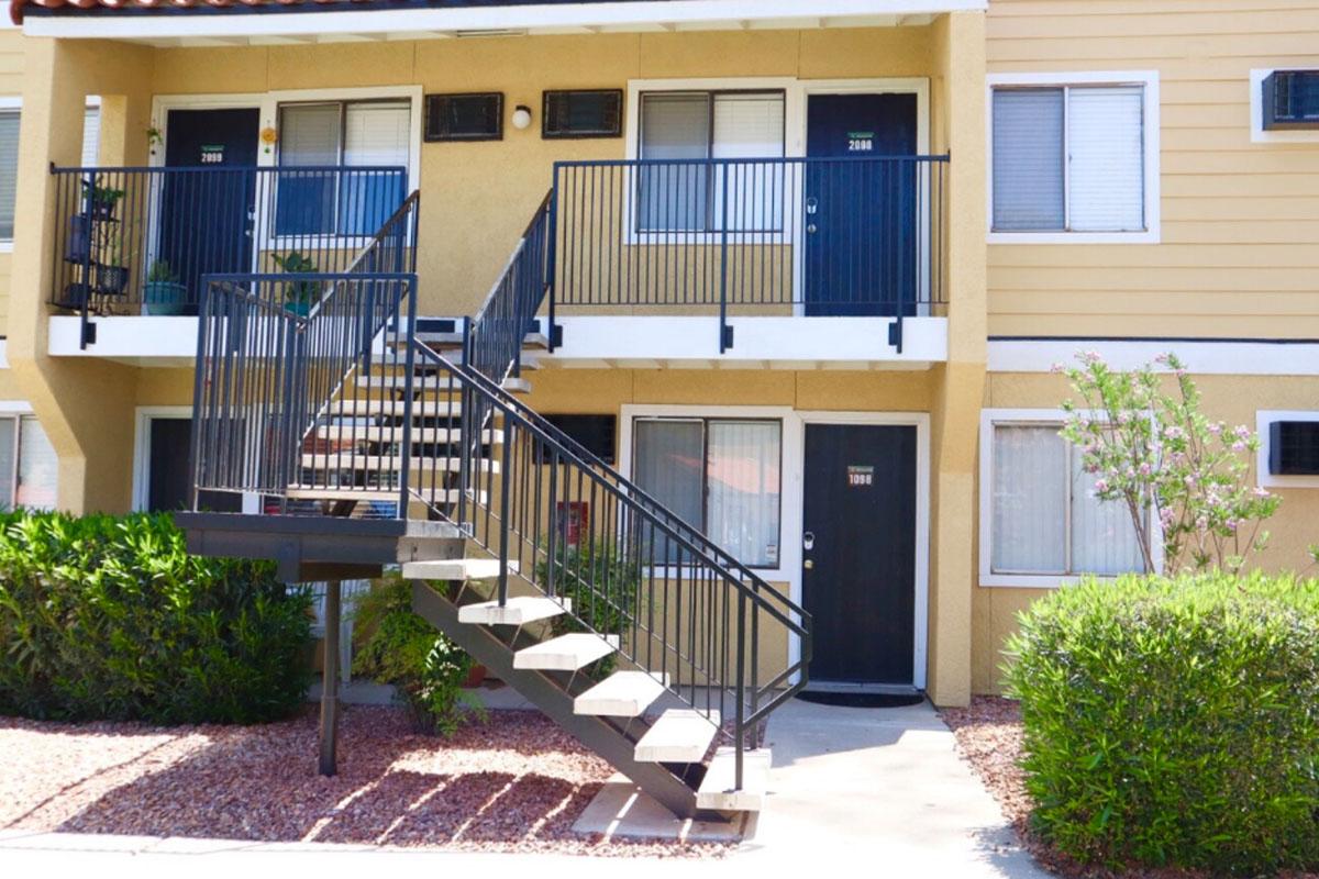 A multi-unit apartment building with two levels. The lower level features two doors, while a staircase leads to the upper floor where two more doors are visible. Neatly landscaped shrubs are in the foreground, and the building is painted in light colors with a textured exterior.