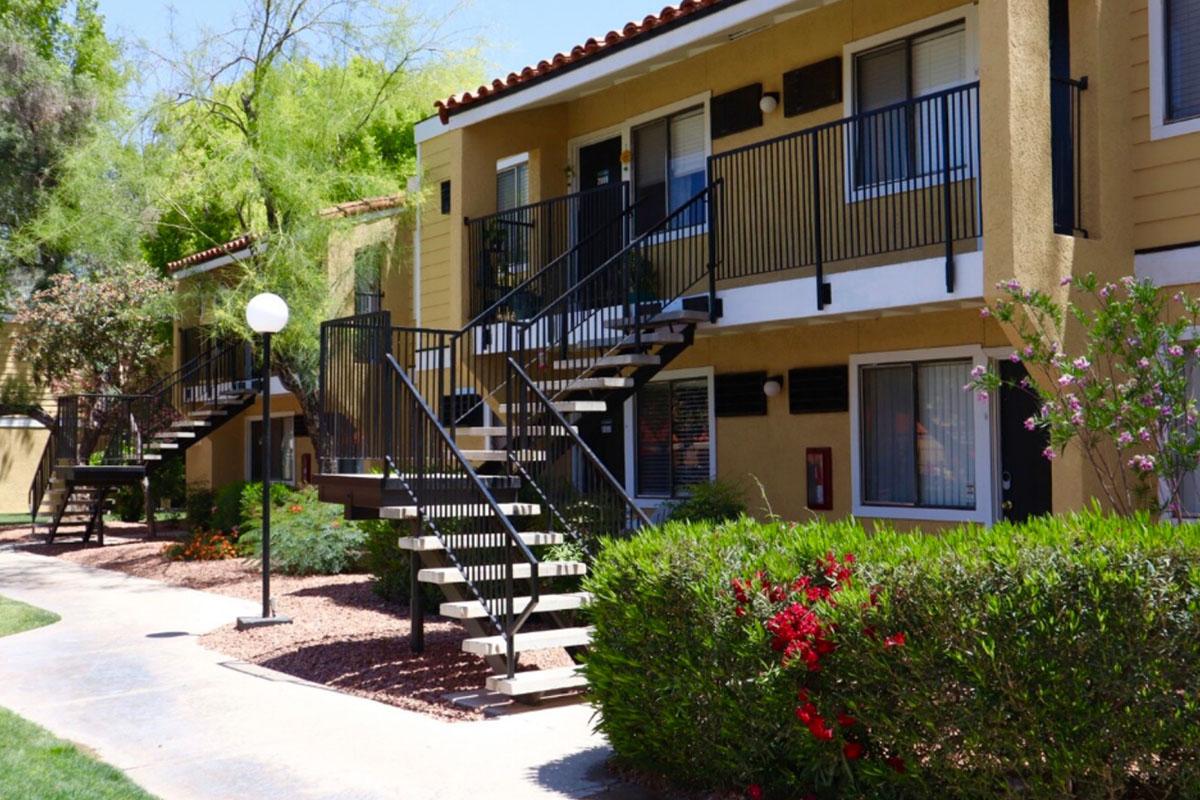 Two-story apartment buildings with outdoor staircases, surrounded by landscaped greenery and colorful flower beds. The pathway leads through the greenery, and a lamppost is visible in front of the buildings. Sunlight shines on the scene, creating a warm, inviting atmosphere.