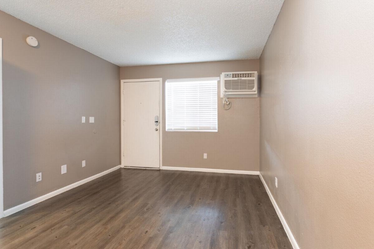 Empty interior of a small room with light brown walls and new flooring. The room features a window with white blinds and an air conditioning unit mounted above it. A door is visible on the right side, while there are wall switches and outlets. The space is well-lit and looks freshly painted.
