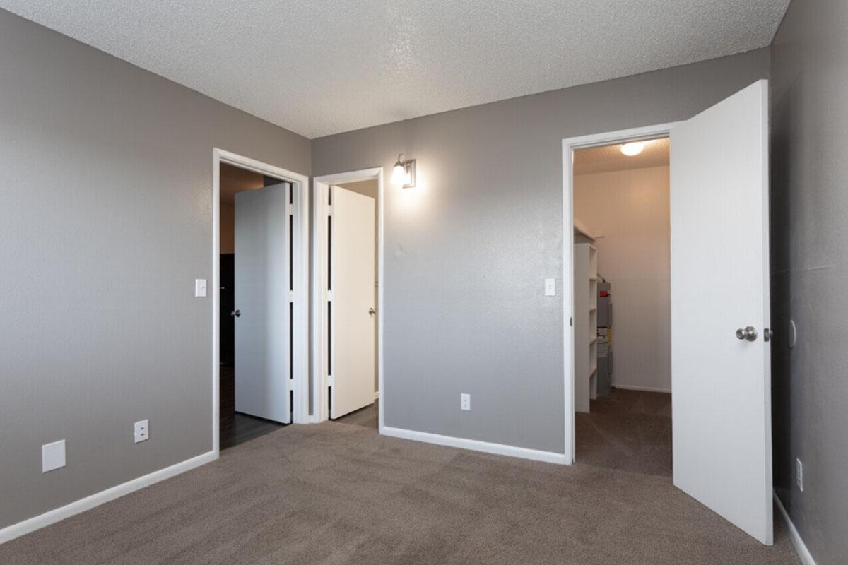 A spacious room featuring light gray walls and carpet. Two white doors are open, leading to adjacent rooms, while a closet door is visible. Soft lighting from a wall sconce adds warmth to the space, creating a clean and modern atmosphere suitable for various uses.