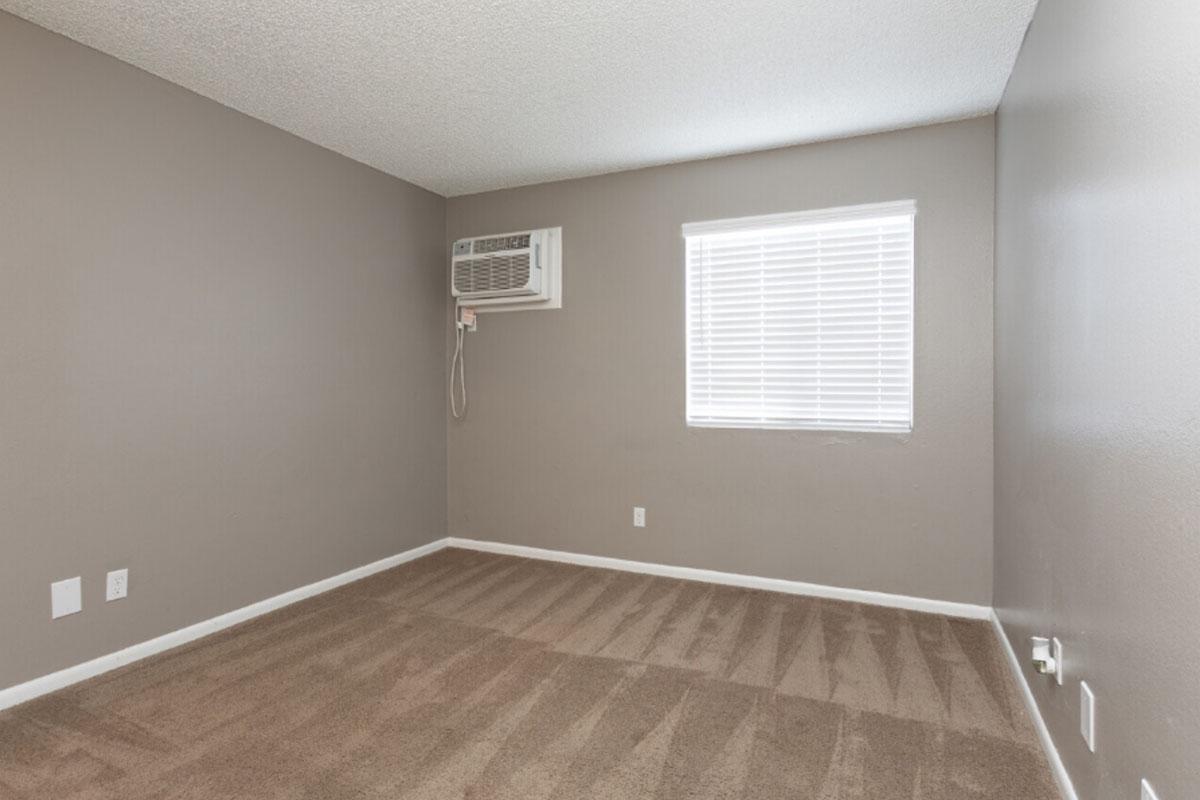 Empty room with light brown carpet and beige walls. An air conditioning unit is mounted on the wall in the corner, and a window with white blinds allows natural light to enter. The space is unfurnished, creating a clean and simple aesthetic.