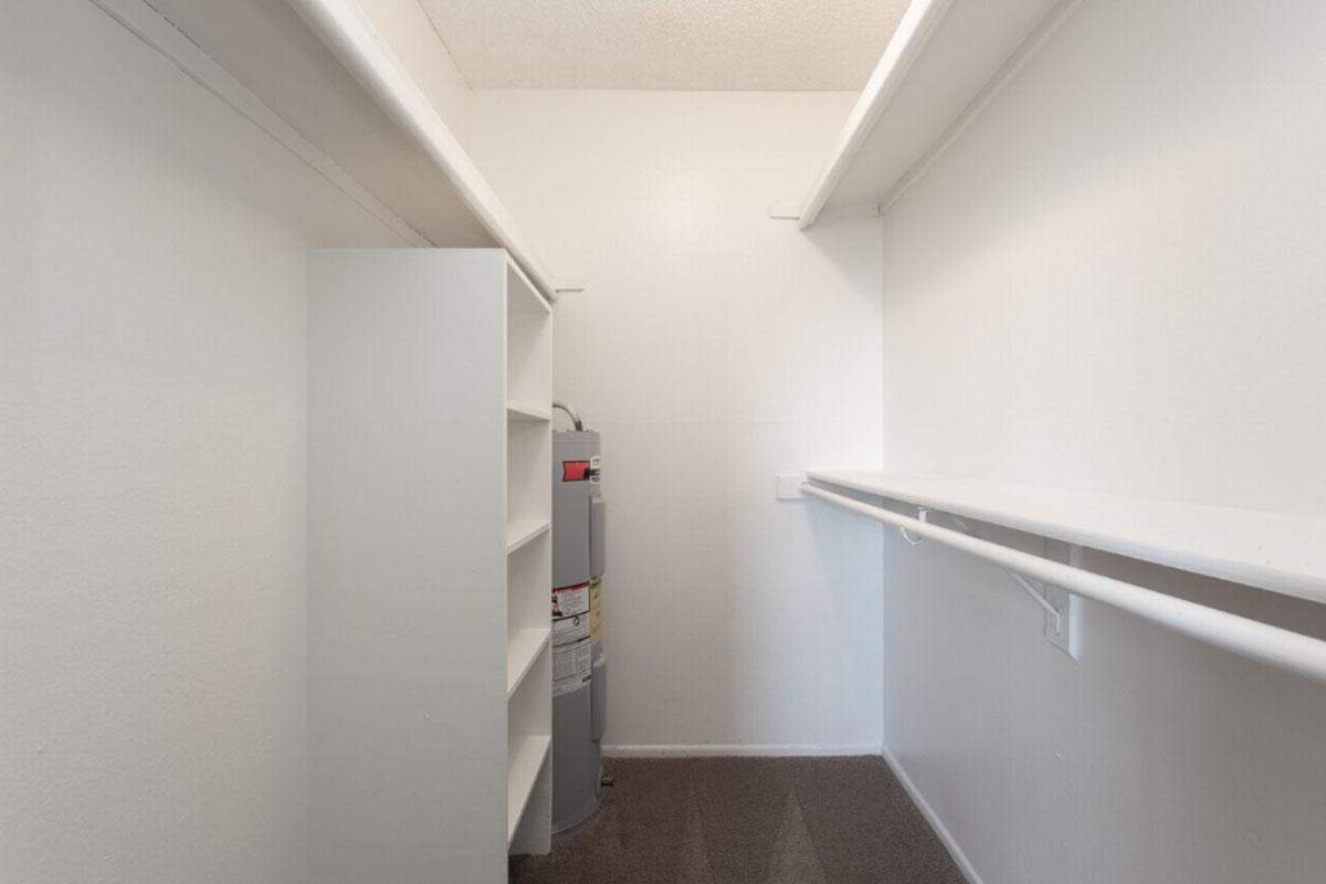 A spacious, empty closet featuring white shelves on one side, a water heater placed against the wall, and a single hanging rod. The walls are painted white, and the floor is covered with carpet, giving it a clean and well-lit appearance.