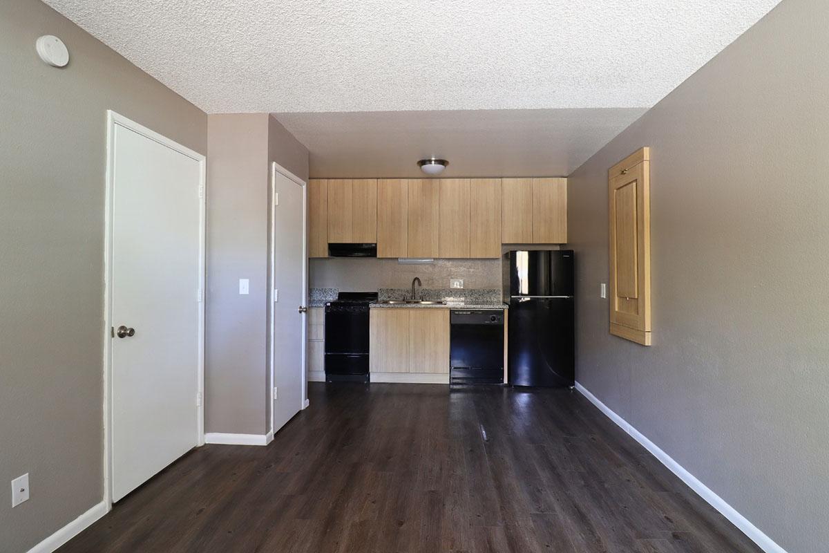 A small, modern kitchen area with wood cabinets and granite countertops, featuring a black refrigerator, stove, and sink. The room has light brown walls and dark wood flooring, with a door leading to another room. A small window is present, and the overall space is well-lit and tidy.