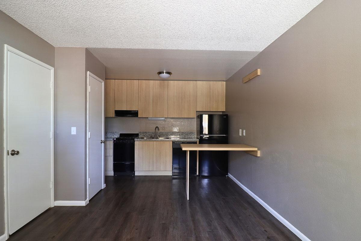 A small kitchen area featuring light wooden cabinets, a black stove, and refrigerator. There's a countertop with a modern sink, and a small fold-out table attached to the wall. The walls are painted a neutral color, and the floor is a dark wood laminate. Natural light is coming from a window out of view.