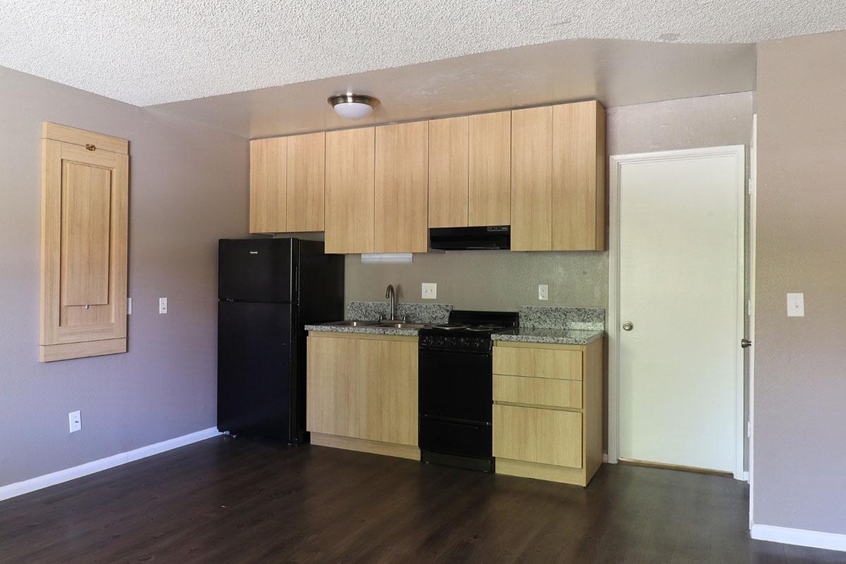 A modern kitchen with wooden cabinetry, black appliances including a refrigerator and stove, and a granite countertop. The walls are painted in a neutral color, and there is a door leading to another area. The flooring is dark, creating a contemporary look.