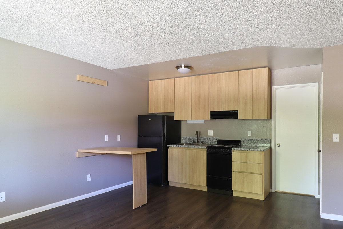 A small kitchen area featuring light wood cabinets, a black refrigerator, and a black stove. There is a gray countertop with minimal decorations and a wall-mounted light fixture. The floor is dark wood, and the walls are painted a neutral color, creating a simple and modern look.