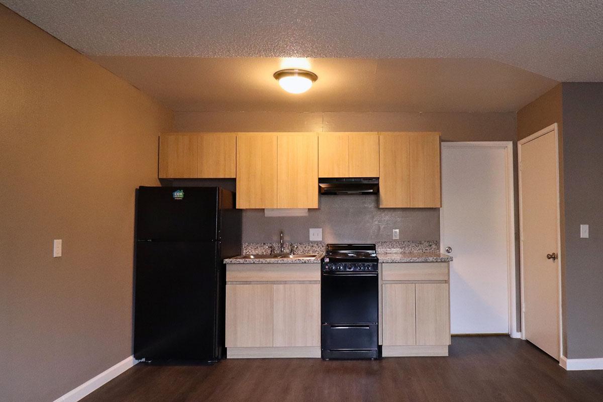 A modern kitchen featuring wooden cabinetry, a countertop with a sink, a black gas stove, and a black refrigerator. The walls are painted gray, and there is a light fixture on the ceiling. A door leads to another room, with a wooden floor completing the space.