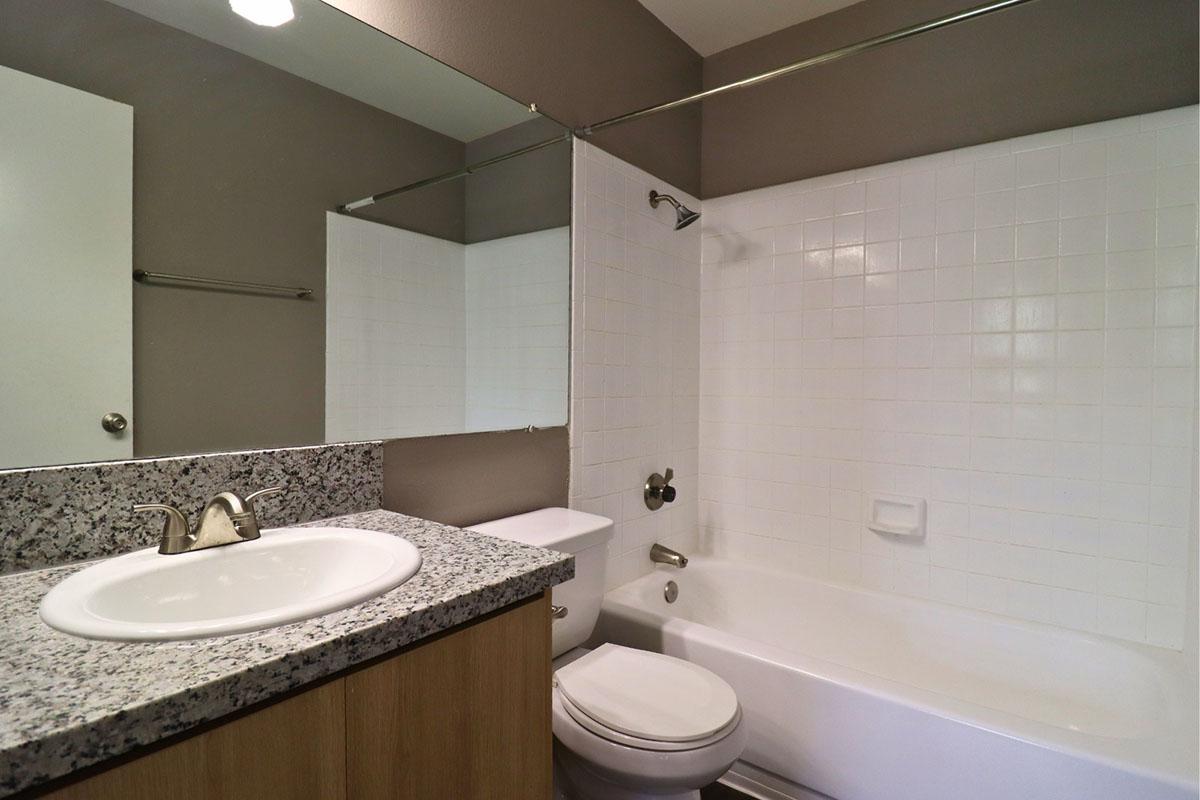 A clean and modern bathroom featuring a white bathtub, a granite countertop sink, a mirror, and a white tiled wall. The space is well-lit with neutral-colored walls and a simple, minimalistic design, creating a fresh and inviting atmosphere.