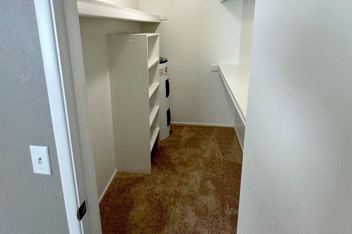 A small, empty room with beige carpet, featuring a white shelf on the left side and a clean, white wall. There's a small appliance or heater against the wall on the right, and a shelf extending along the far wall. The space appears tidy and well-lit, with a doorframe visible on the left.