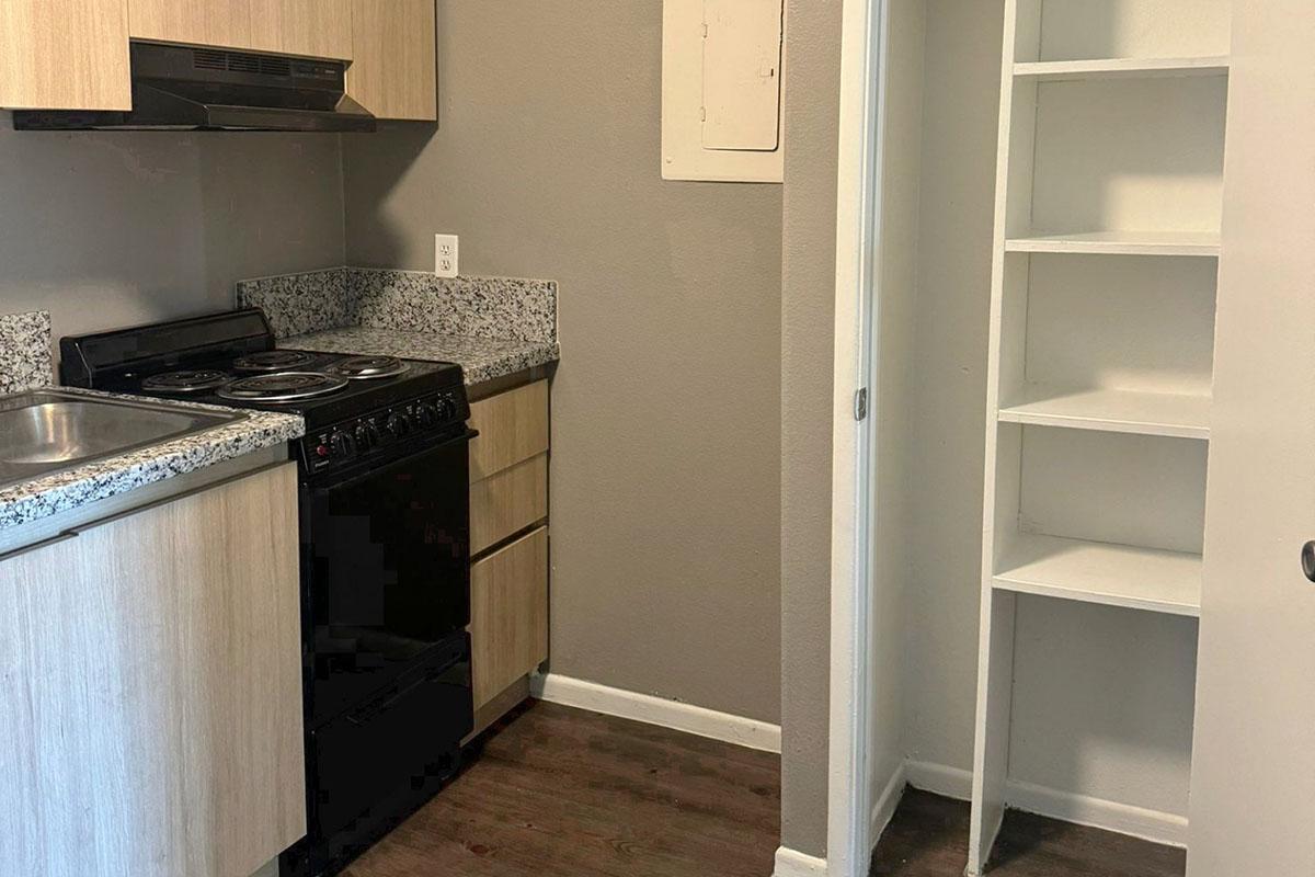 A compact kitchen featuring a stove, oven, and sink built into wooden cabinetry with a granite countertop. Next to the kitchen area, there is a small pantry with white shelving and a door that opens to it. The walls are painted gray, and the floor is dark wood.