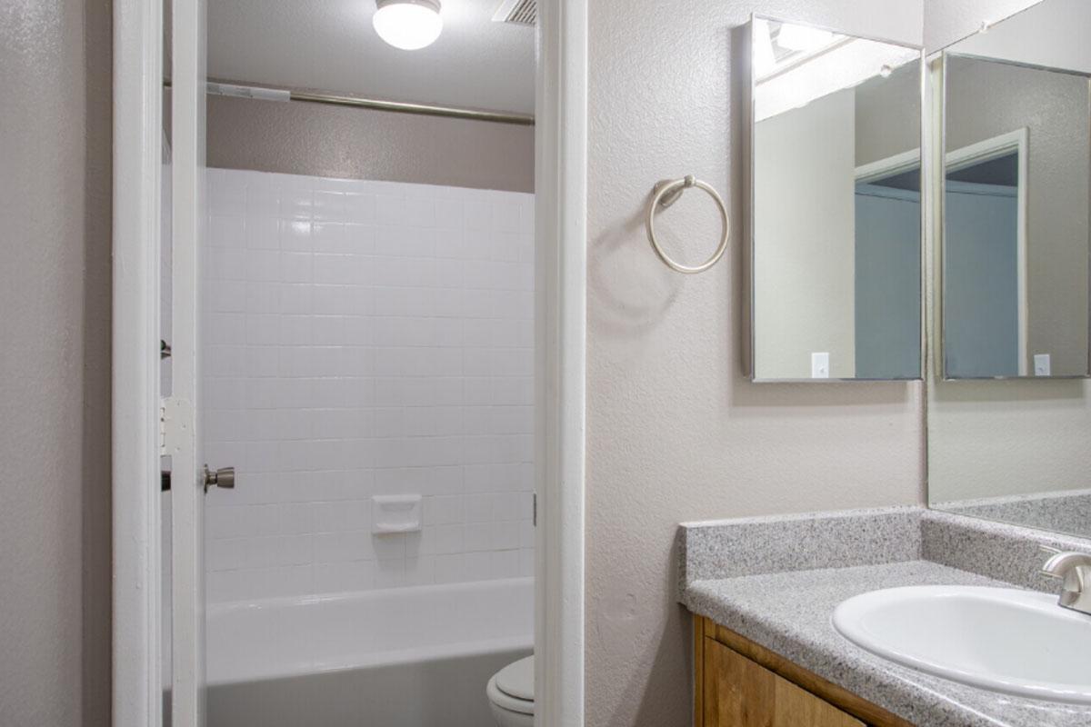 A clean bathroom interior featuring a white tiled bathtub, a single sink with a countertop, a mirror above the sink, and another mirror on the wall. The lighting is bright, and the walls are painted in a light color, enhancing the spacious feel of the area. A towel ring is mounted on the wall.