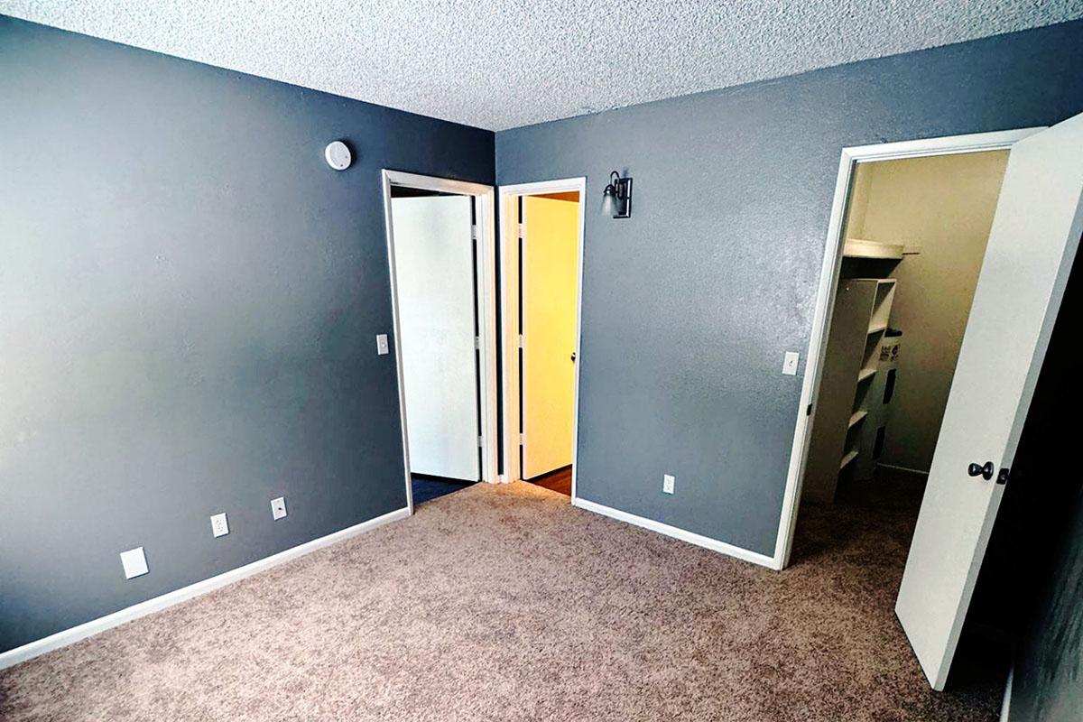 A view of a small, empty room with gray walls and carpeted flooring. There are two doors visible: one leading to a closet area and another to an adjacent room. A light fixture is mounted on one wall, and there are several electrical outlets visible. The space appears clean and well-lit.