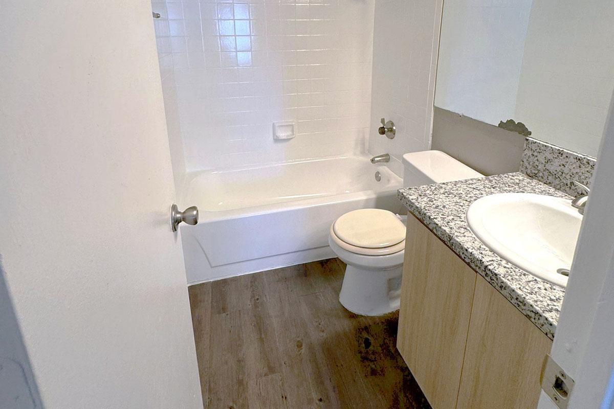 A clean, modern bathroom featuring a white bathtub, a toilet, and a sink with a granite countertop. Light wooden flooring complements the white tiles on the wall, creating a bright and spacious atmosphere. The room has a simple design with neutral colors and minimal decor.