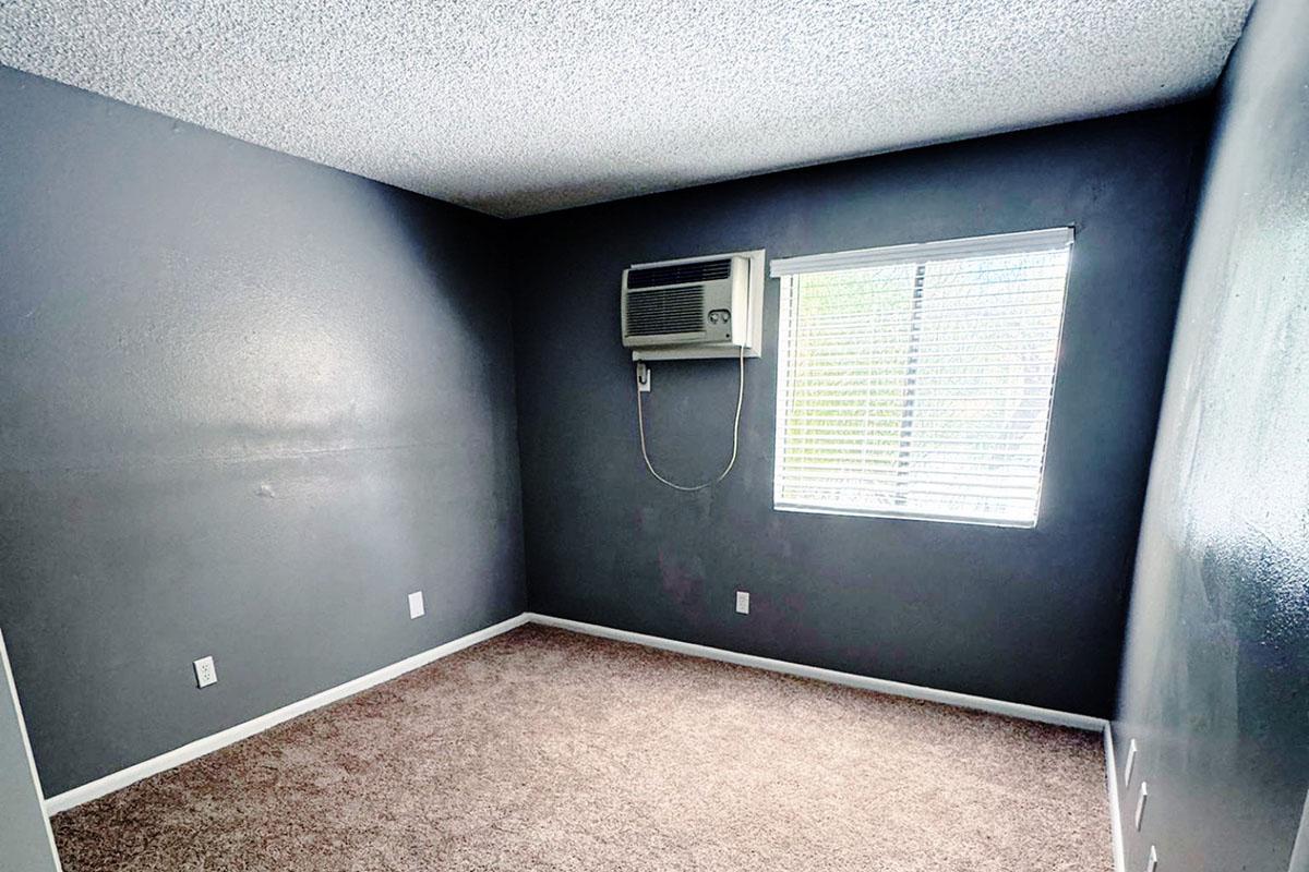 Empty room with dark gray walls and carpeted flooring. An air conditioning unit is mounted on the wall near a window with blinds. The room is sparsely furnished, showcasing a simple and minimalist design. Natural light filters in through the window, illuminating the space.