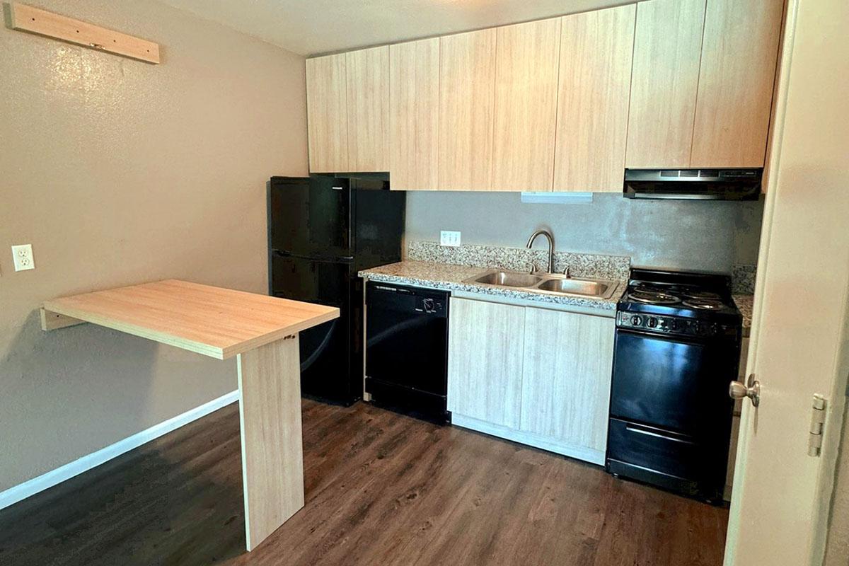 A small kitchen featuring wooden cabinetry, a granite countertop, and modern appliances including a gas stove, a black refrigerator, and a black dishwasher. There is also a compact wooden table attached to the wall. The walls are painted in a light gray color, and the flooring is dark wood.