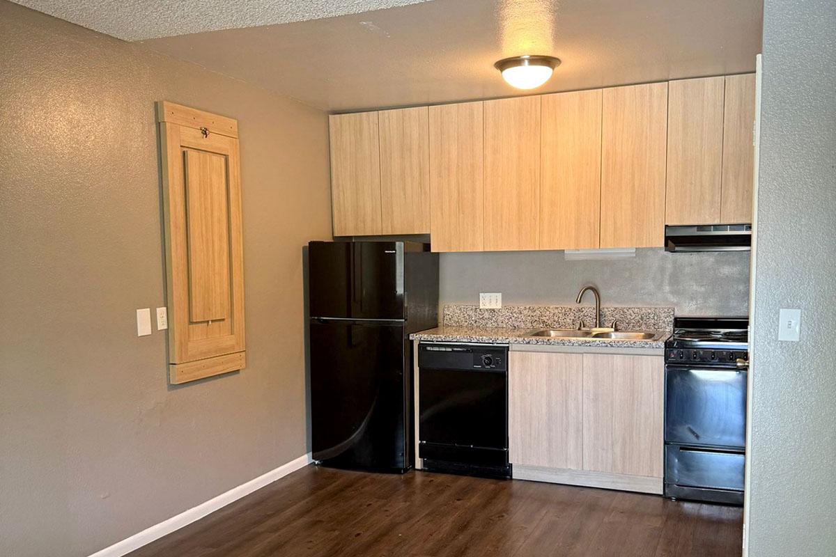 Modern kitchen featuring light wood cabinetry, a black refrigerator, and a black dishwasher. The countertop is made of speckled granite. There is a stove and an overhead microwave. The walls are painted a muted gray, and the floor has dark wood laminate. A sliding cabinet door is visible on the left.