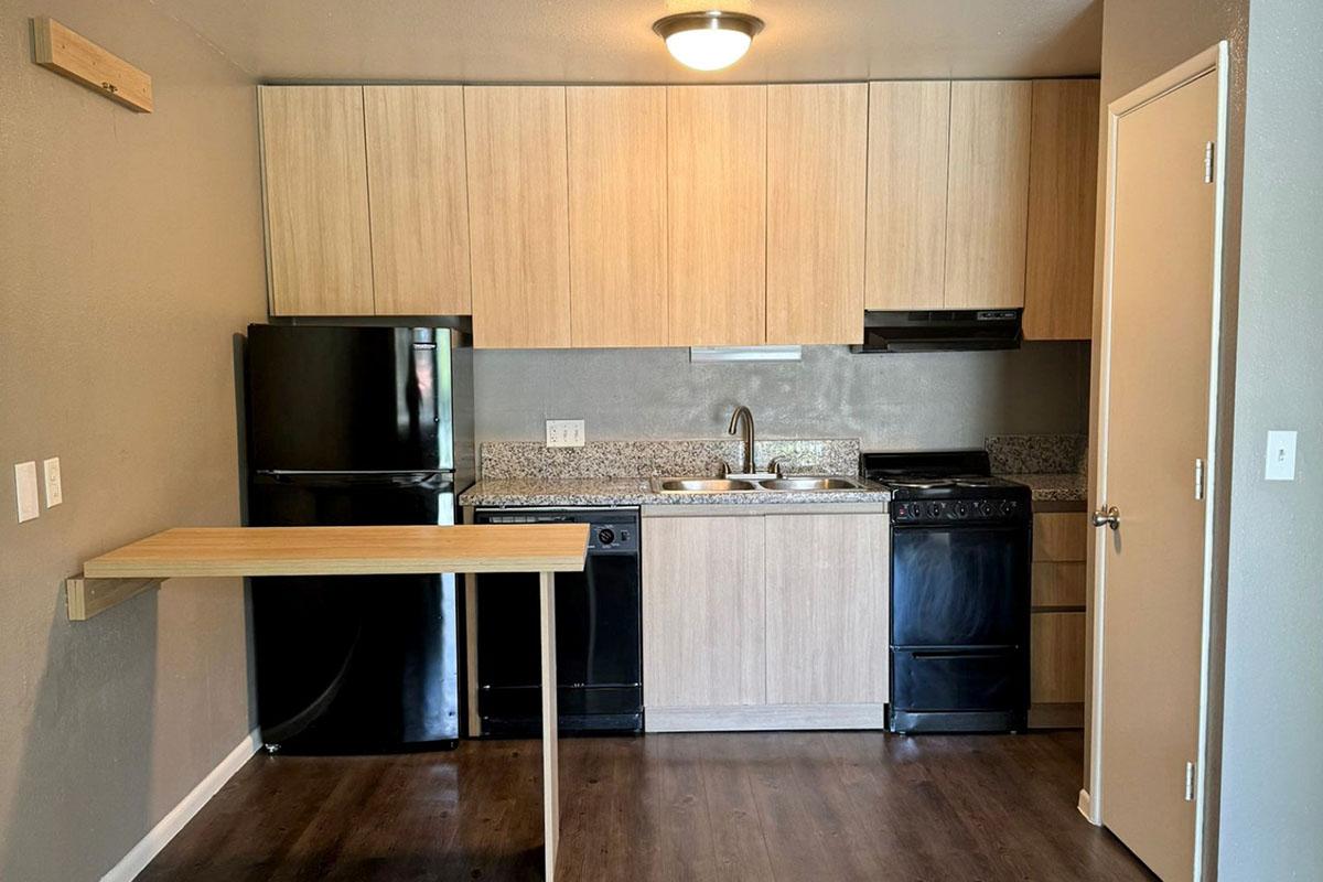 A modern kitchen featuring light wooden cabinets, a black refrigerator, dishwasher, and stove. The countertop is a speckled gray design. A small, minimalist table is positioned against the wall, and the room is painted in neutral tones with a door leading to another area.