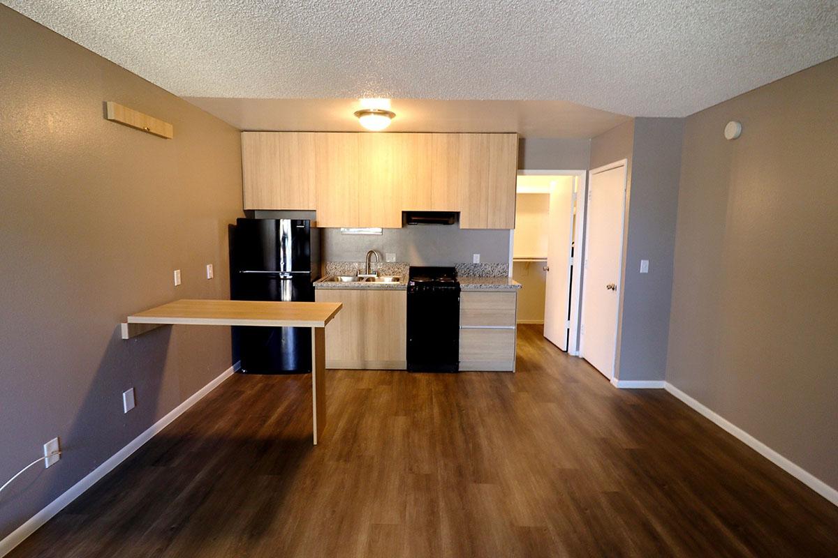 A modern kitchen and living area featuring a countertop with a small table, dark refrigerator, and wooden cabinetry. The space has neutral-colored walls and wood-like flooring, with a doorway leading to another room visible in the background. The lighting is bright, creating a welcoming atmosphere.