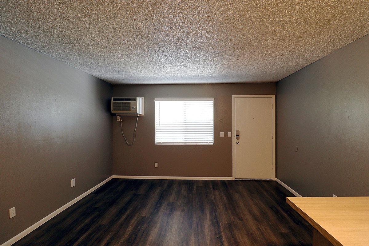 A spacious, empty room with a textured ceiling, featuring a window with horizontal blinds on one wall. An air conditioning unit is mounted above the window, and a front door is visible on the right. The flooring is dark wood laminate, with light brown walls creating a neutral backdrop.