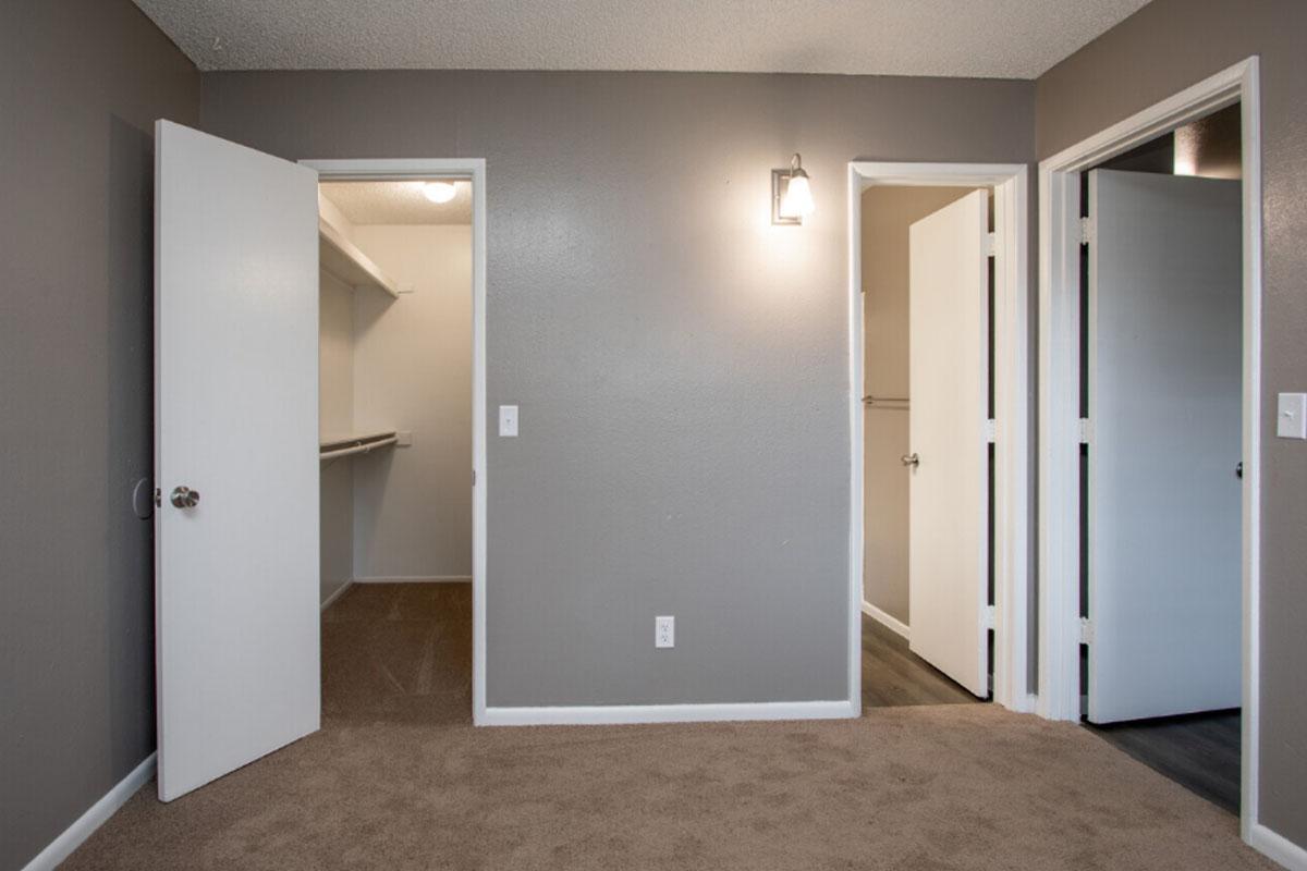 A small room with light brown carpet and gray walls. Two doors on the right lead to closets or other spaces, while a third door opens to a closet with shelves. A wall-mounted light fixture provides illumination. The room appears tidy and well-maintained.