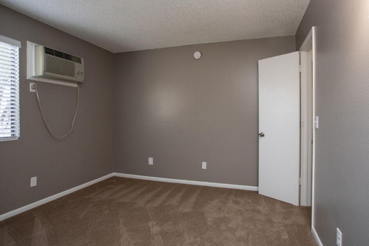 A small, empty room featuring beige carpet and gray walls. There is a wall-mounted air conditioning unit on the left, a small round vent on the ceiling, and a closed white door on the right. A window with blinds is visible, allowing natural light to enter.