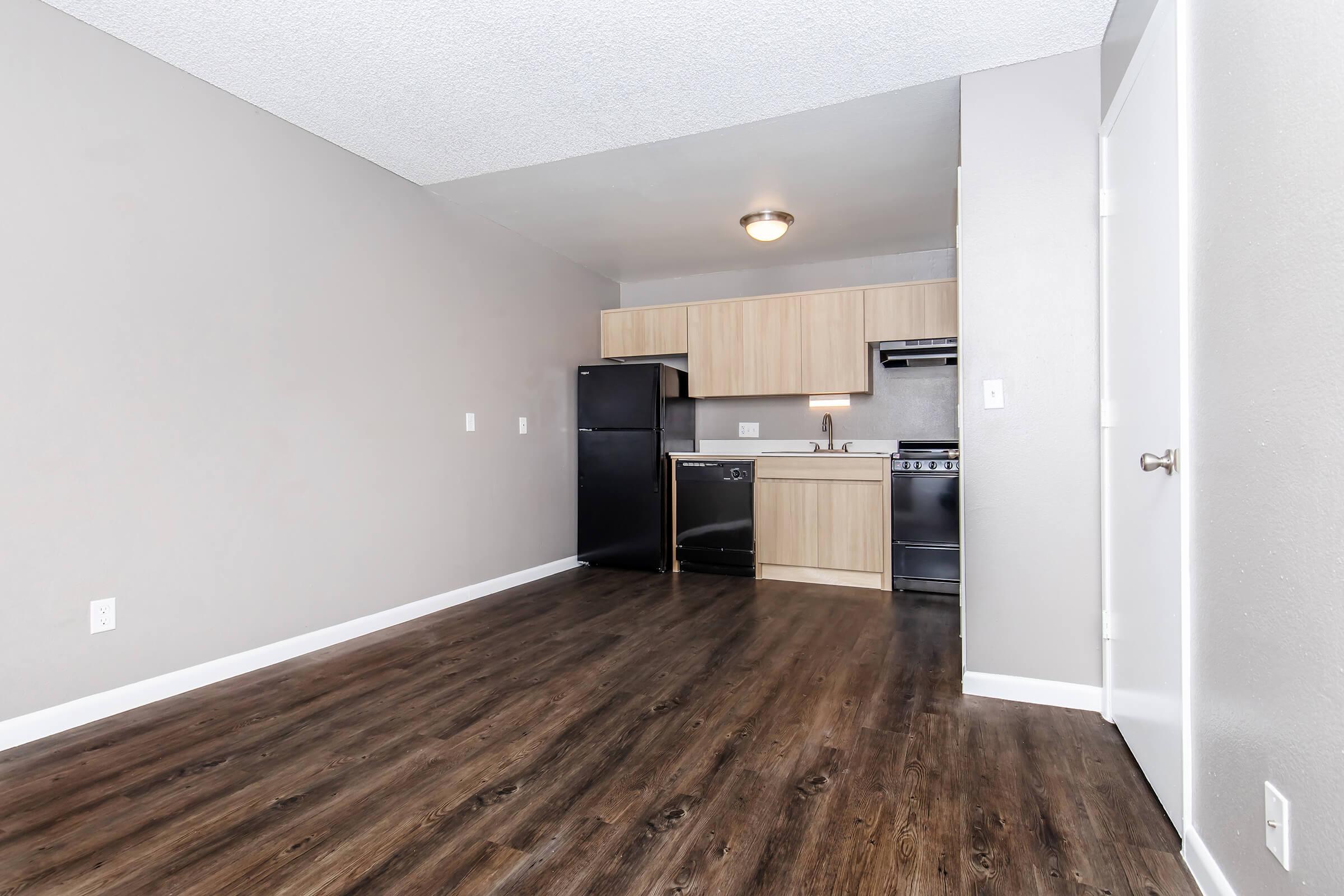 A modern kitchen area in a residential apartment featuring wood-like flooring, beige walls, and light wood cabinetry. Appliances include a black refrigerator, a stove, and a dishwasher. The open space has a ceiling light, creating a bright and inviting atmosphere.