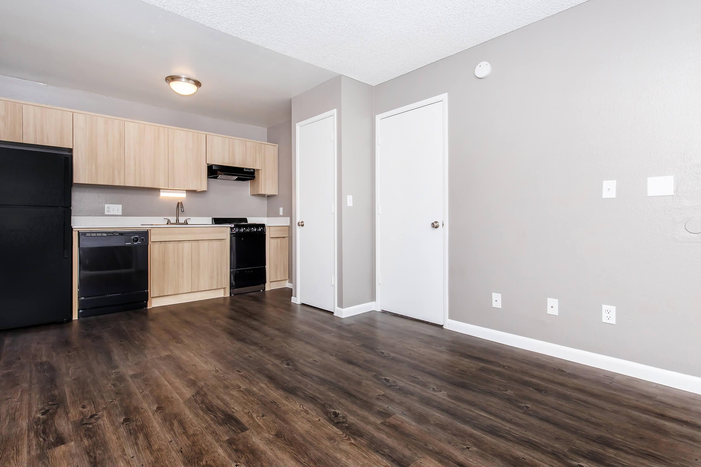 A modern kitchen area with light wooden cabinetry, black appliances, and an open floor plan. The space features a dark laminate wood floor and neutral gray walls, with doors leading to other rooms. A ceiling light fixture provides illumination.