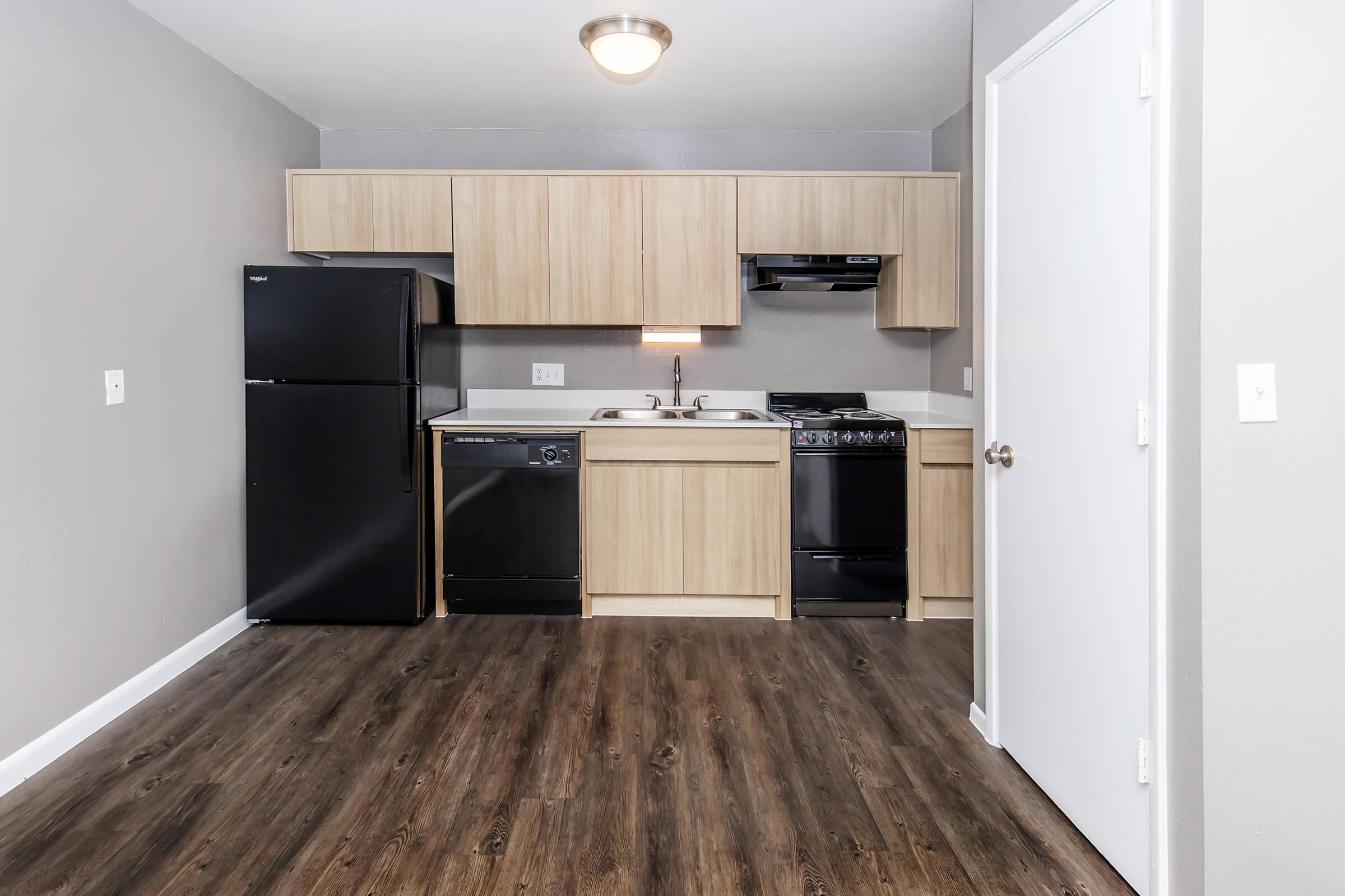 Interior view of a modern kitchen featuring wood cabinets and appliances. The kitchen includes a black refrigerator, dishwasher, and stove. The countertop is white, and the floor is a dark wood laminate, creating a sleek and contemporary look. Walls are painted in a light gray color.