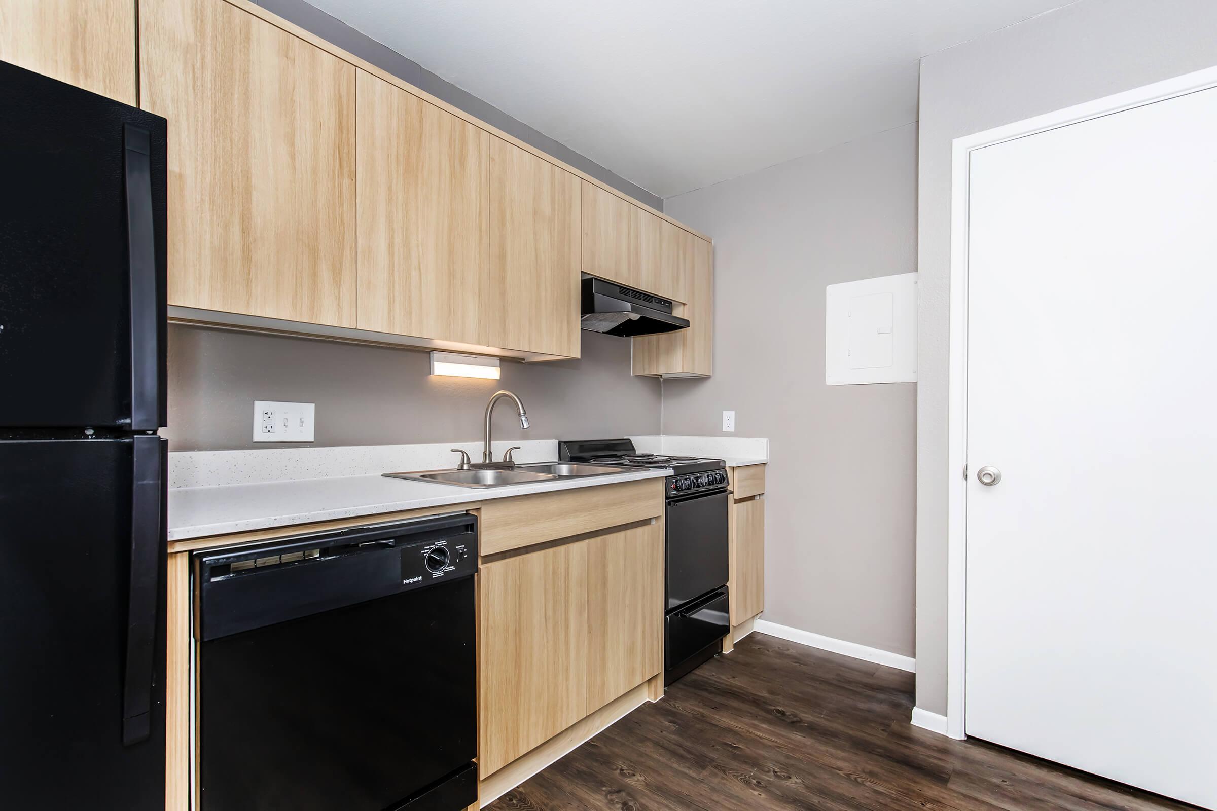 Modern kitchen featuring light wood cabinetry, a black refrigerator, a black dishwasher, and a stainless steel sink. The countertop is white, and the walls are painted a neutral gray. A door leads to another room, enhancing the open layout of the space.