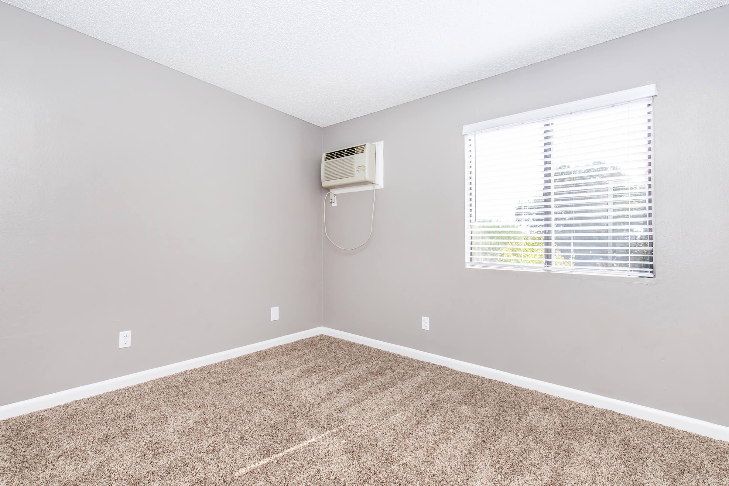 A spacious, empty room with light gray walls and a carpeted floor. An air conditioning unit is mounted on the wall, and there is a window with blinds allowing natural light to enter. The overall atmosphere is bright and clean, suggesting it is ready for furnishing or decoration.