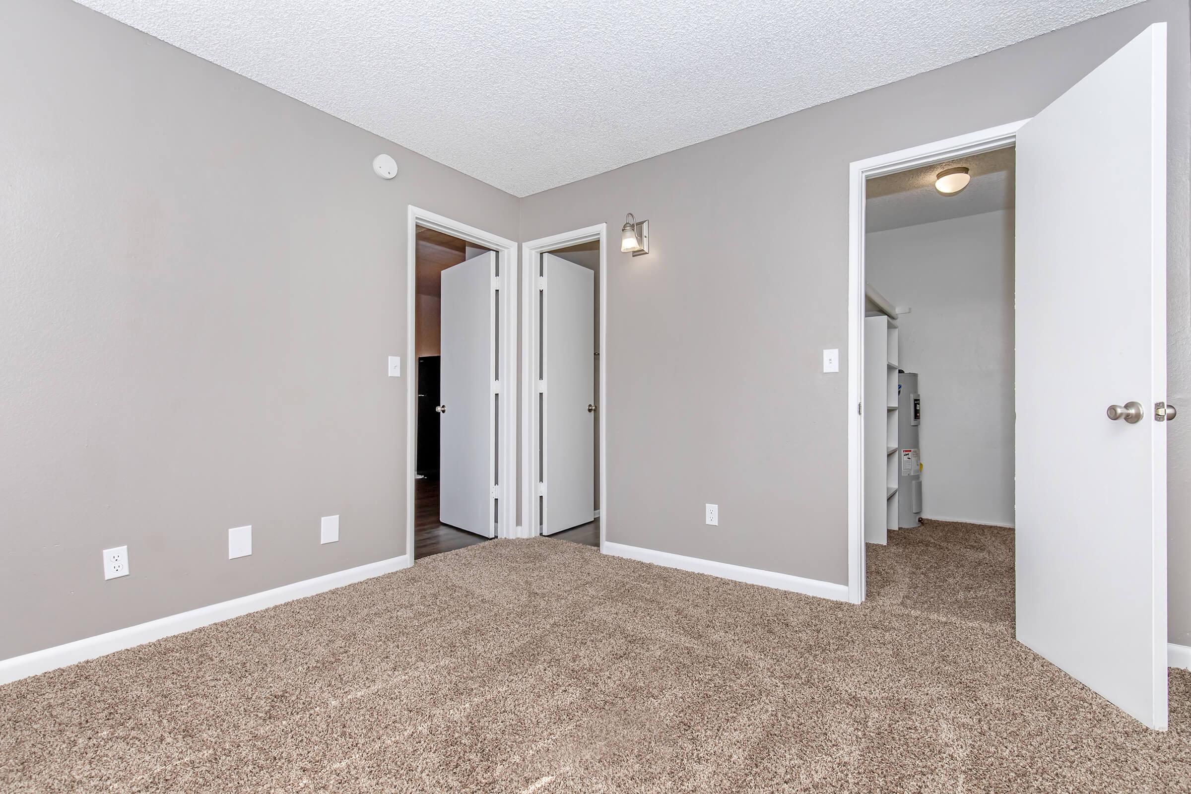 A light gray room with soft carpet, featuring two open white doors leading to other spaces. A wall light fixture is mounted on the left wall. The room appears spacious and well-lit, with a clean and modern aesthetic.