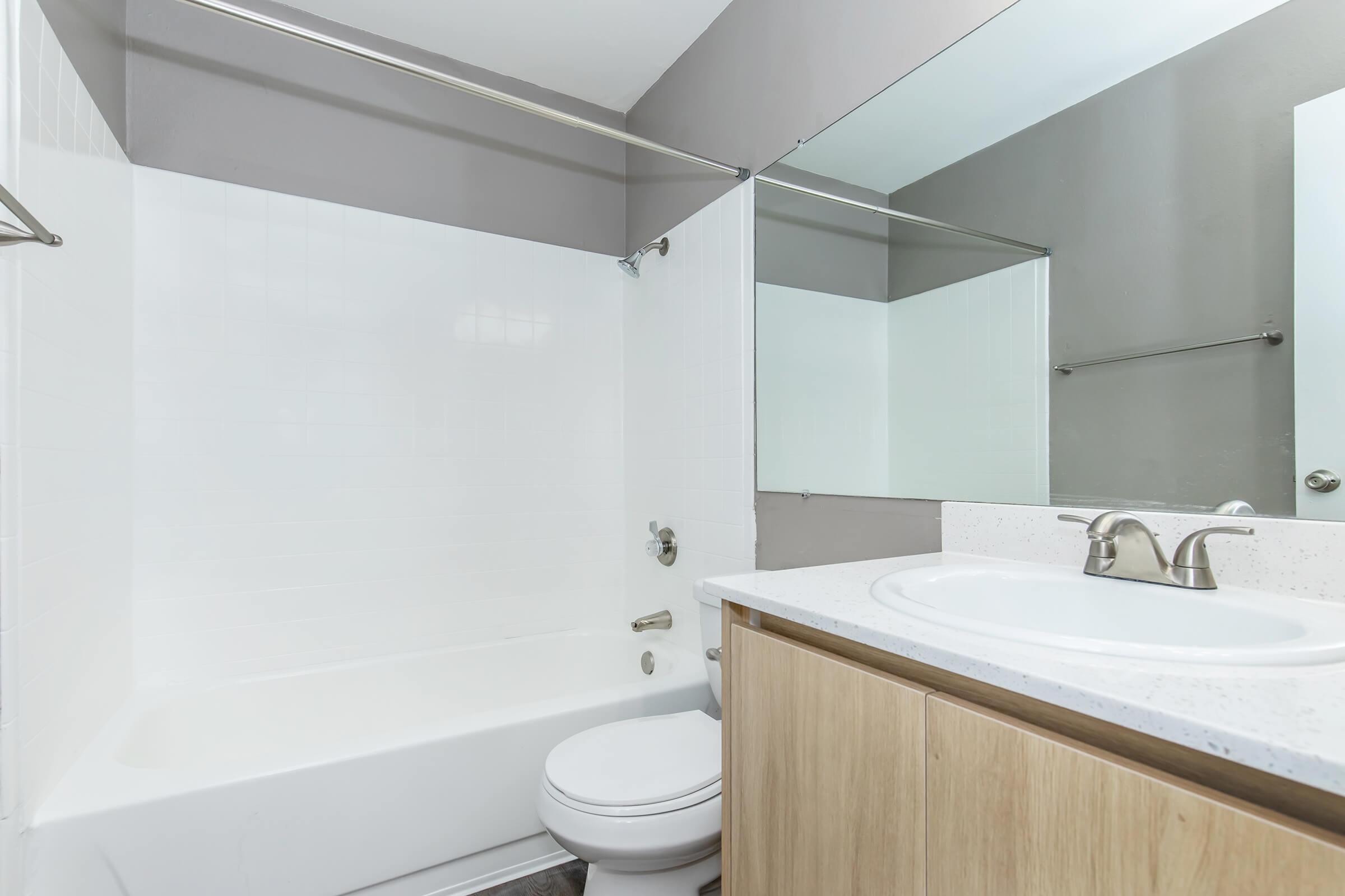 A modern bathroom featuring a white bathtub with a shower, a clean countertop with a sink, and a mirror above. The walls are painted in a light gray color, and there are wooden cabinets underneath the sink. The overall design is contemporary and minimalistic.