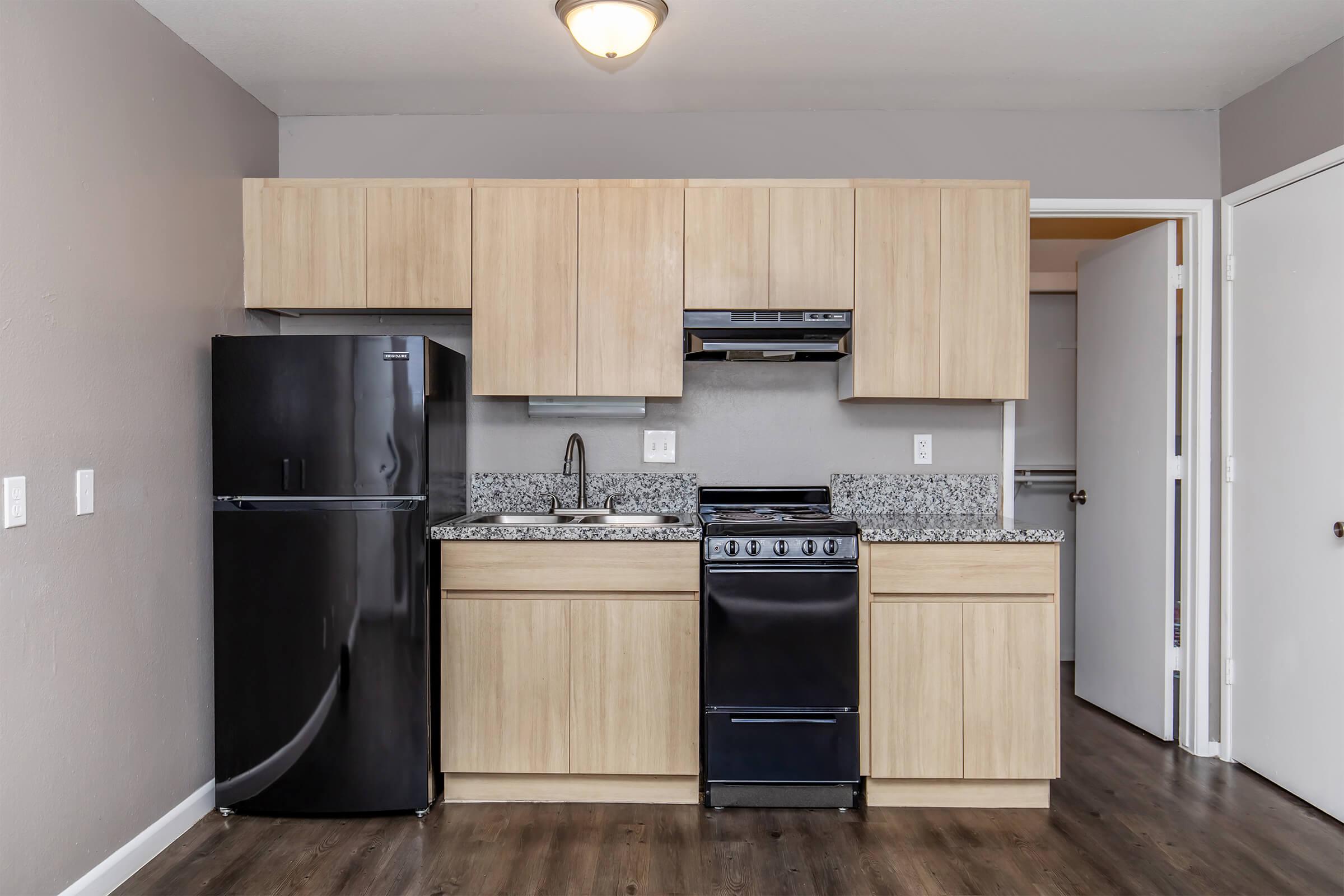 Modern kitchen featuring light wood cabinets, a black refrigerator, a black stove, and a granite countertop. The space has gray walls and wooden flooring, with a doorway visible leading to another room.