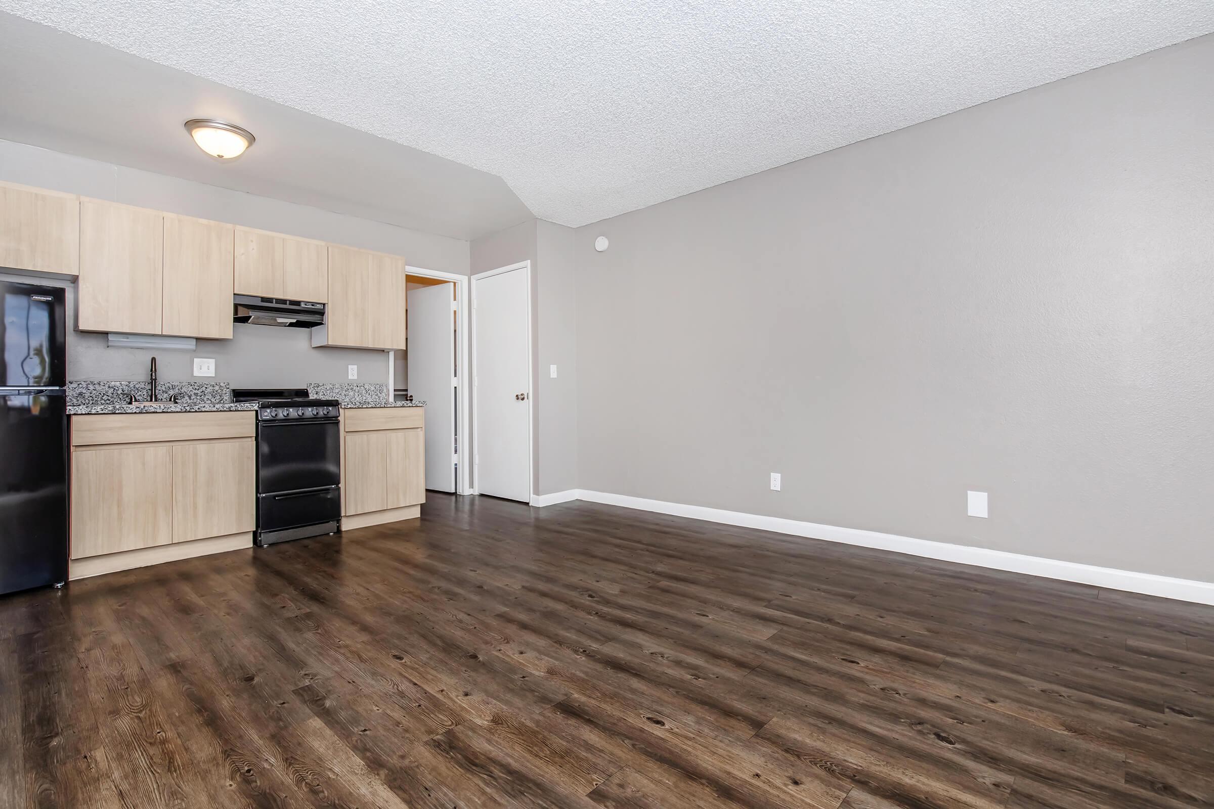 Interior view of a modern kitchen and living area. The kitchen features light-colored wood cabinets, black appliances, and granite countertops. The living area has hardwood-style flooring and neutral gray walls. A door leads to another room, while the space is bright and open.