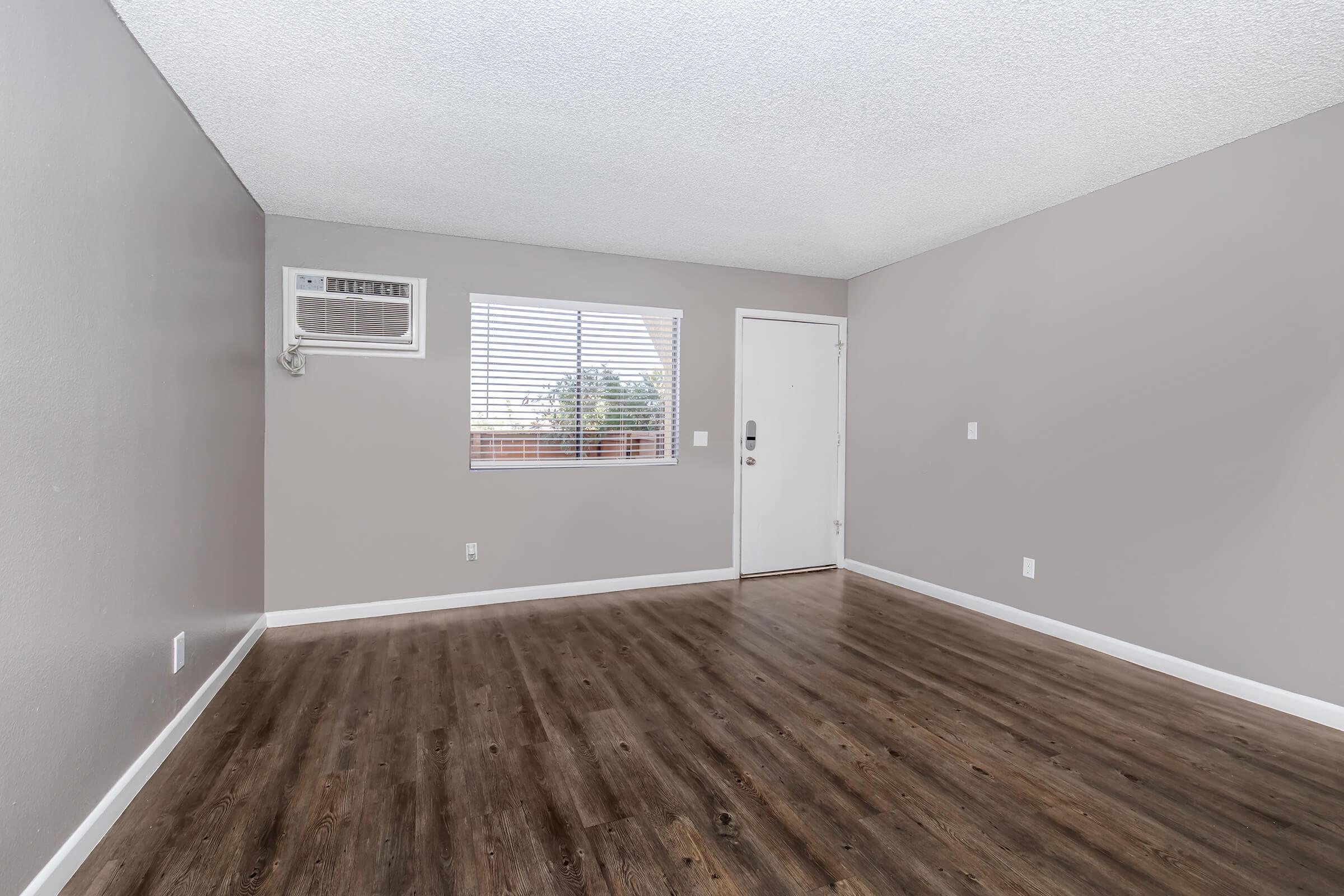 Empty room with light gray walls and a wood-like floor. A window with blinds allows natural light, and there's an air conditioning unit on the wall. The doorway leads outside, and the space is devoid of furniture, creating a clean and airy atmosphere.