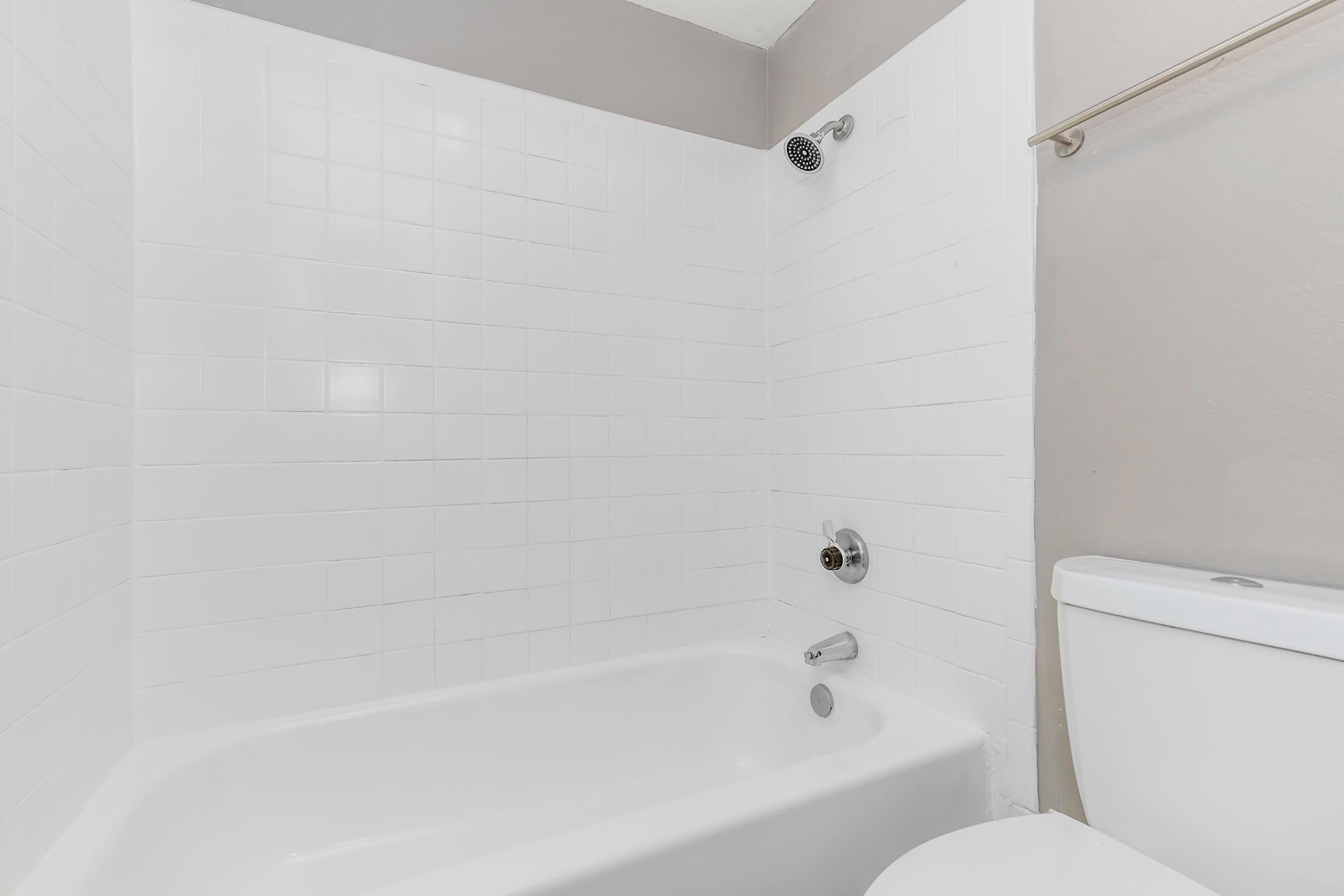 A clean, modern bathroom featuring a white bathtub with a showerhead above, white tiled walls, and a toilet nearby. The space has a simple, minimalist design with neutral colors.