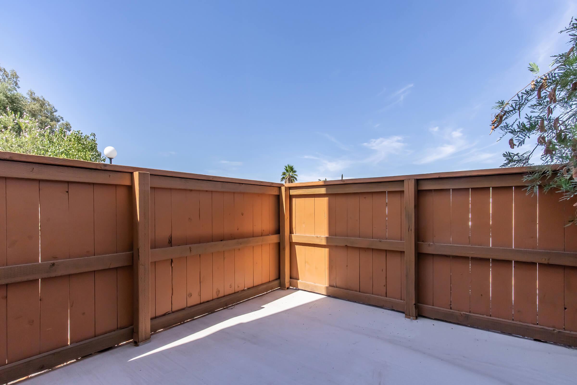 A wooden fence enclosing a small outdoor space, with a clear blue sky above and a few trees visible in the background. The sunlight casts shadows on the ground, creating a serene and bright atmosphere.