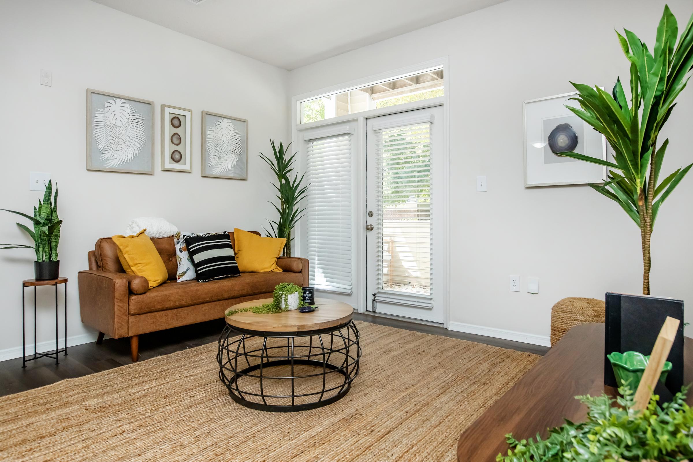 Cozy living room featuring a brown sofa with yellow accents, a round coffee table, and a large rug. There are indoor plants, a decorative wall art, and sliding glass doors leading outside. The space is well-lit with natural light and has a modern, inviting atmosphere.