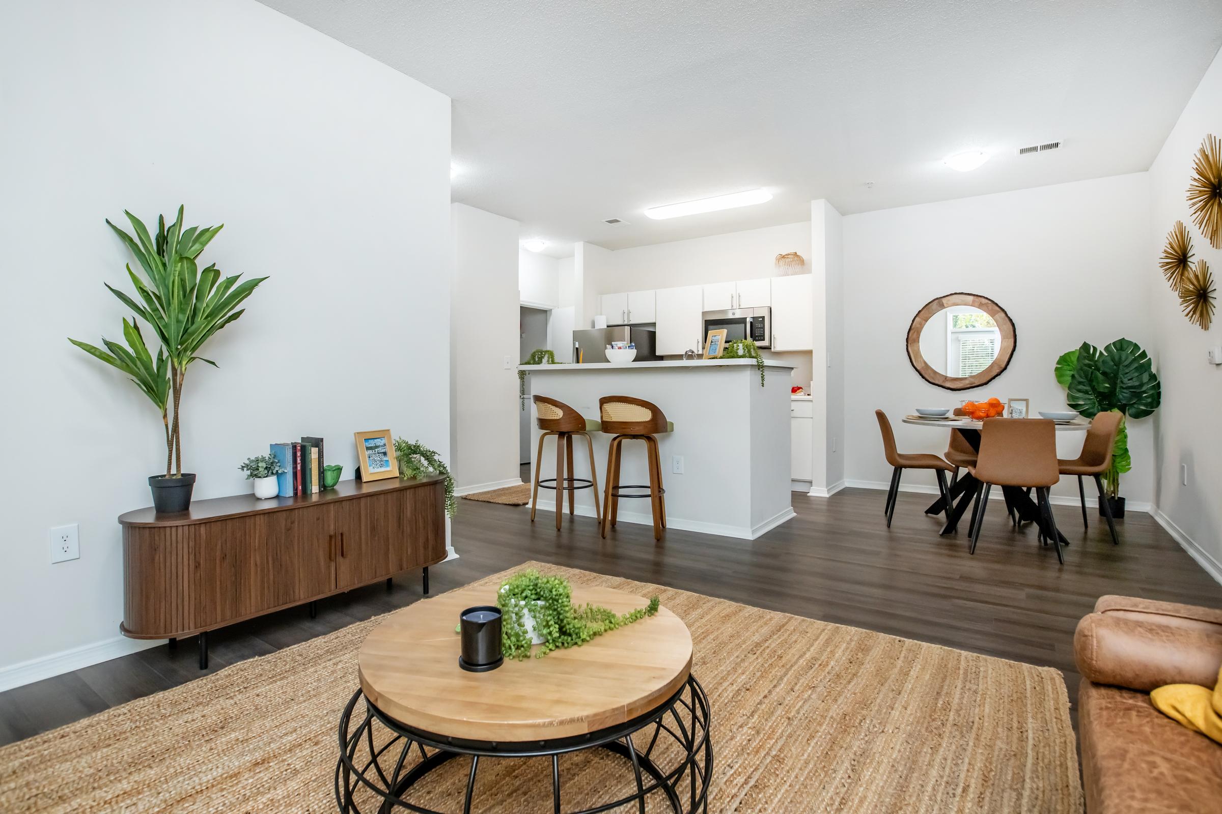 A modern living space featuring a cozy seating area with a brown sofa, a round coffee table, and a decorative rug. In the background, there's a kitchen with white cabinets and bar stools. A dining area with a round table and chairs is visible, along with green plants and wall decor, creating a welcoming atmosphere.