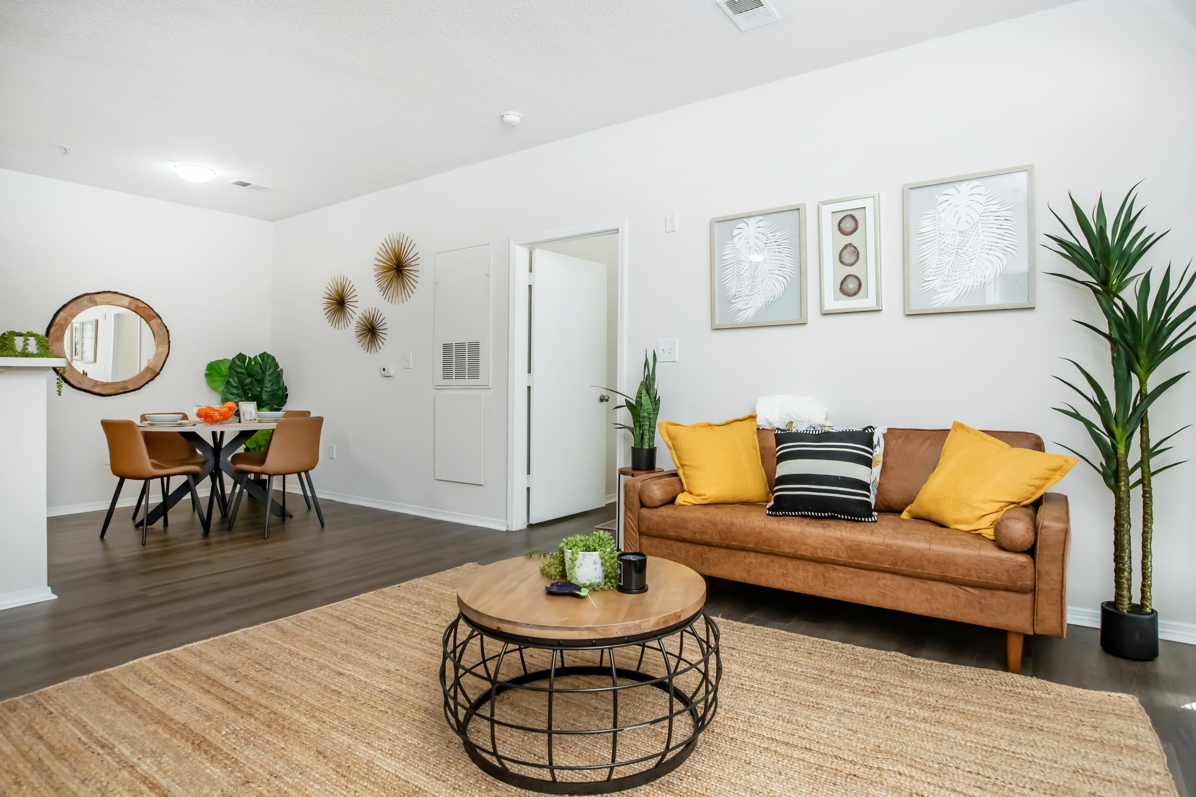 Modern living room featuring a brown leather sofa with yellow and black accent pillows. A round coffee table sits on a textured rug, and there are tropical plants in the corners. In the background, a dining area with a small table and chairs is visible, along with decorative wall art.
