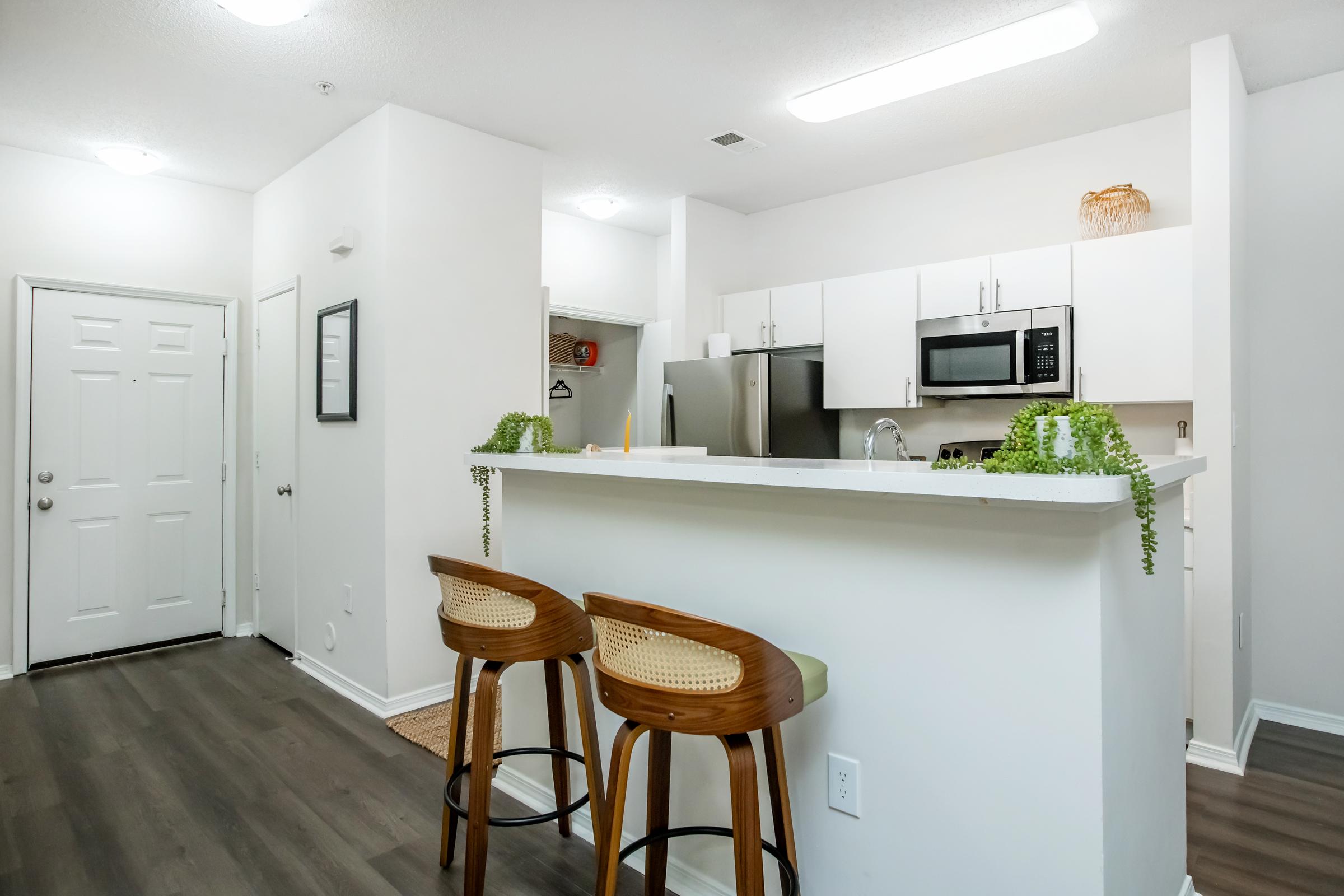 A modern kitchen area featuring a white countertop with two wooden bar stools, stainless steel appliances, and minimalist cabinetry. An open layout connects the kitchen to a light-filled entrance with a door. Bright lighting and plants add a fresh touch to the space.