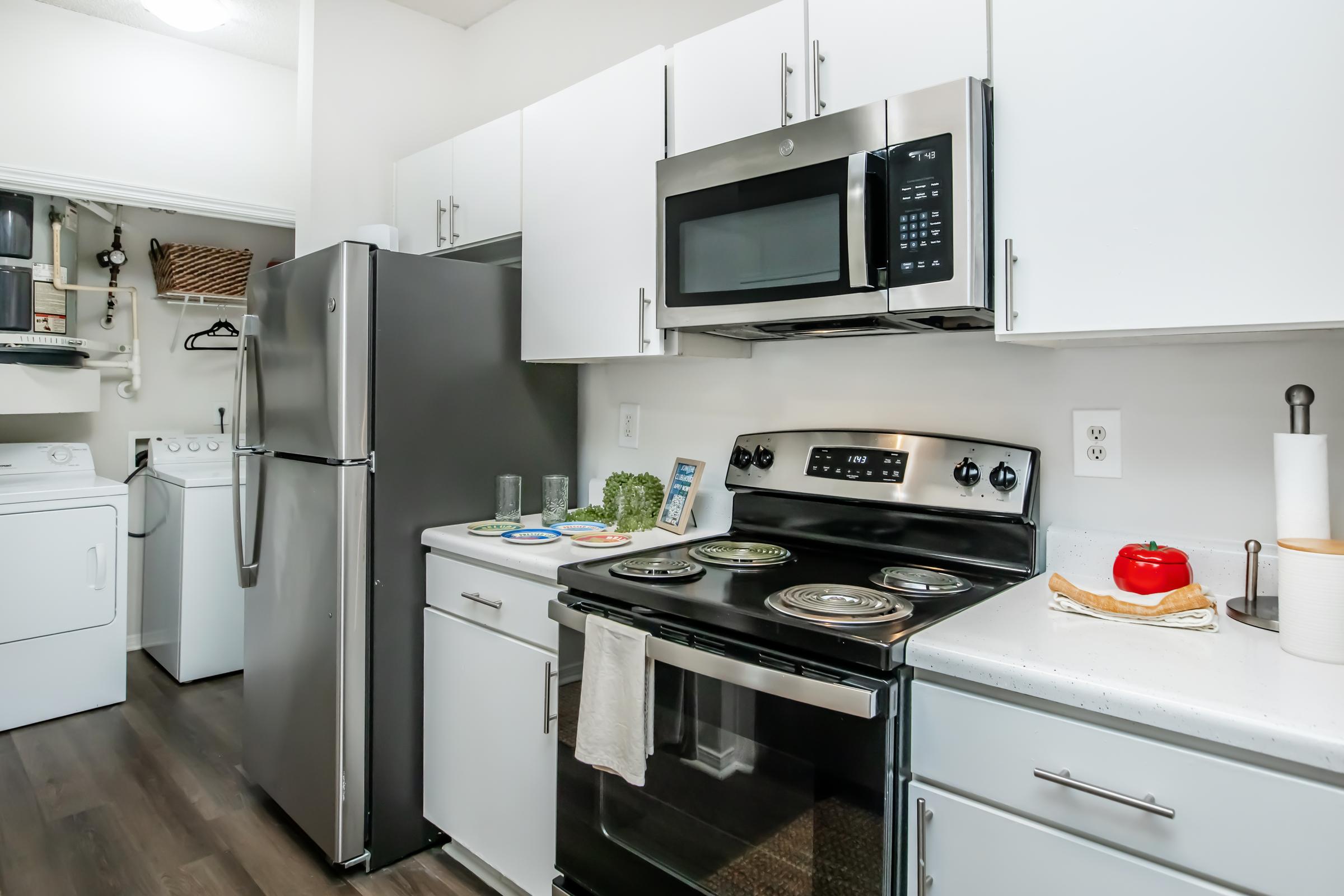 A modern kitchen featuring stainless steel appliances, including a refrigerator and microwave, along with a black stove. The cabinets are white, and there's a blender and decorative items on the counter. In the background, a laundry area with a washer and dryer is visible.