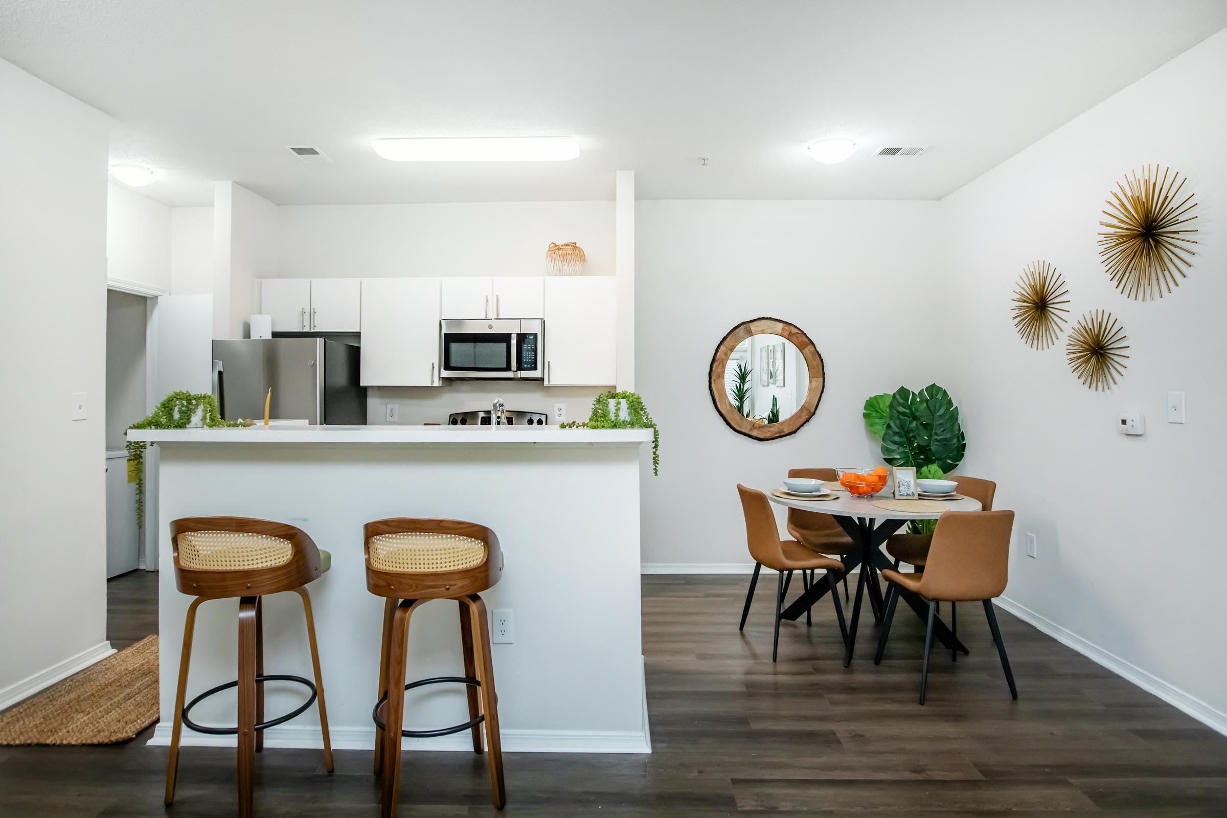 Modern kitchen and dining area featuring a bar with two stools, a round dining table set for four, and decorative wall mirrors. The kitchen includes stainless steel appliances and white cabinetry. Greenery accents add a fresh touch to the minimalist aesthetic. Warm wood flooring enhances the inviting atmosphere.