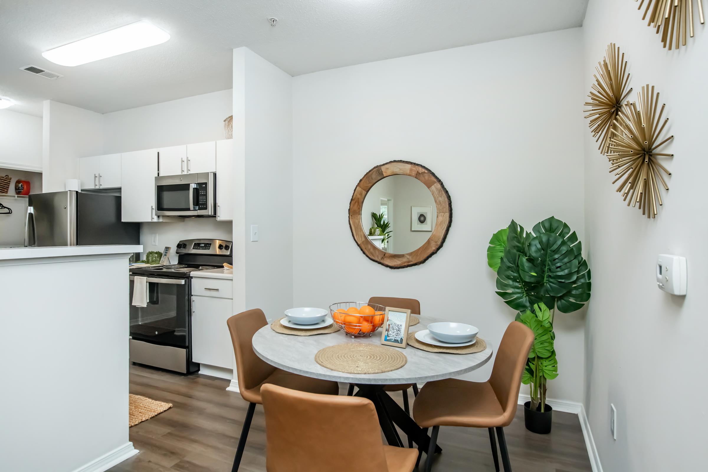 A modern dining area featuring a round table set with plates and a bowl of oranges. Behind it, a kitchen area with stainless steel appliances, and on the wall, a round mirror framed by wood. A large green plant adds a touch of nature, while decorative gold wall pieces enhance the ambiance.
