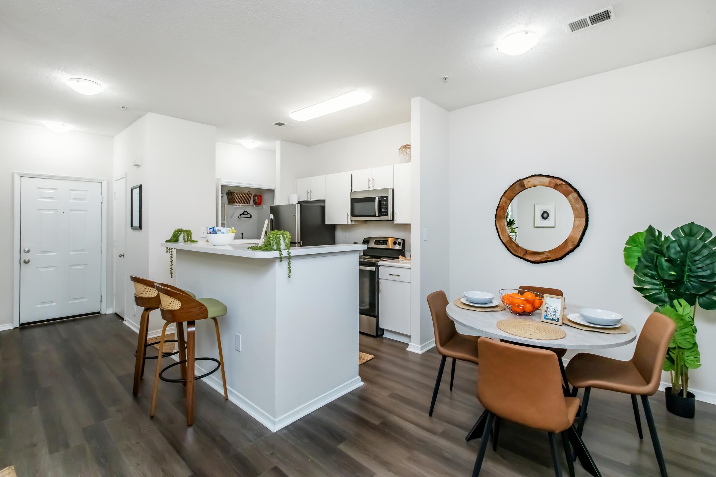 Open-concept kitchen and dining area featuring modern appliances, a round mirror on the wall, a dining table set with plates and an orange centerpiece, and stylish seating. The space is well-lit with natural light and includes potted plants for a fresh look.