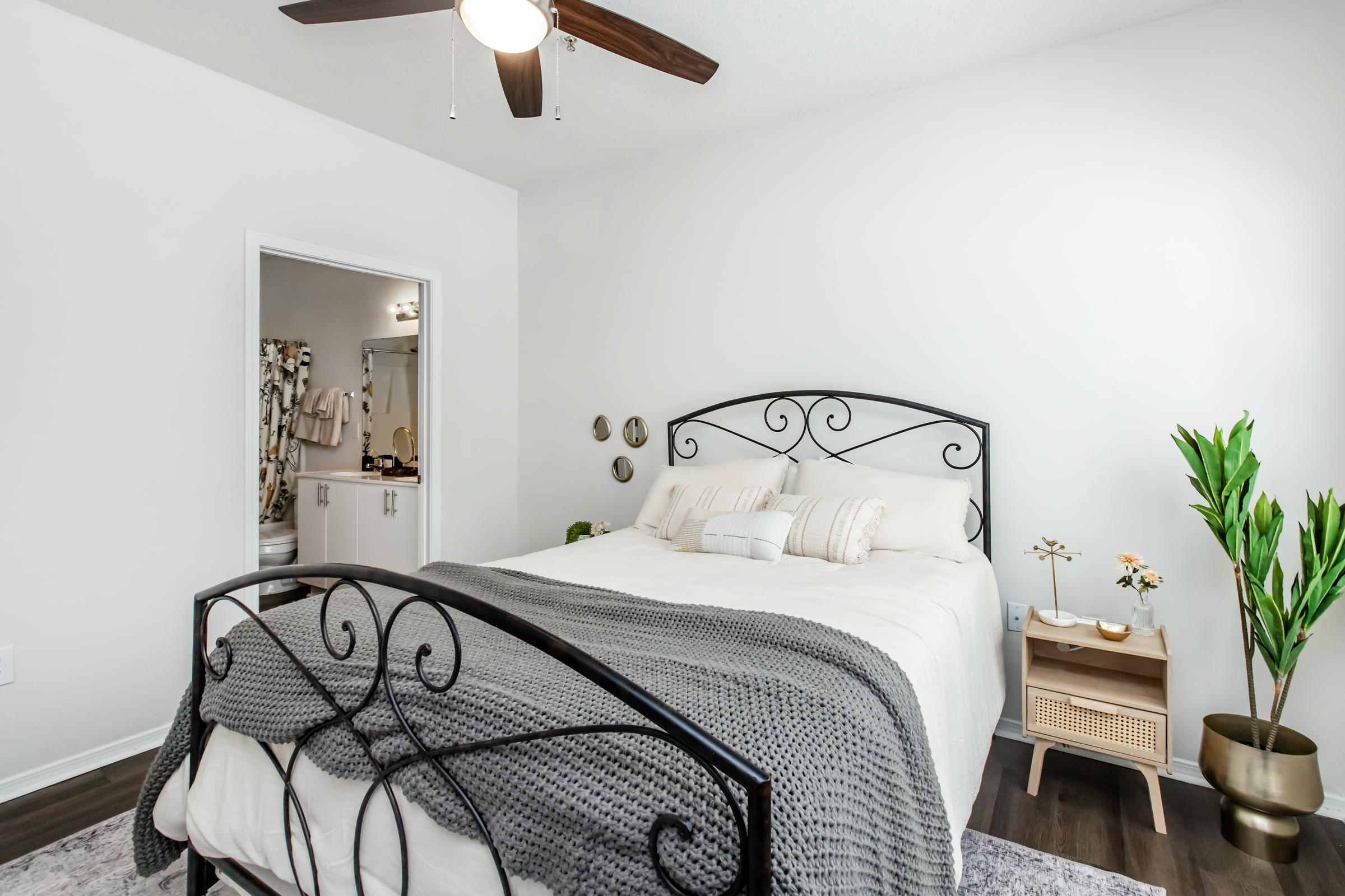 A cozy bedroom featuring a black wrought iron bed with a neutral-colored blanket, two pillows, and a small nightstand with decorative items. A plant adds a touch of greenery, while a bathroom is visible through an open door in the background. The walls are painted white, creating a bright and airy atmosphere.