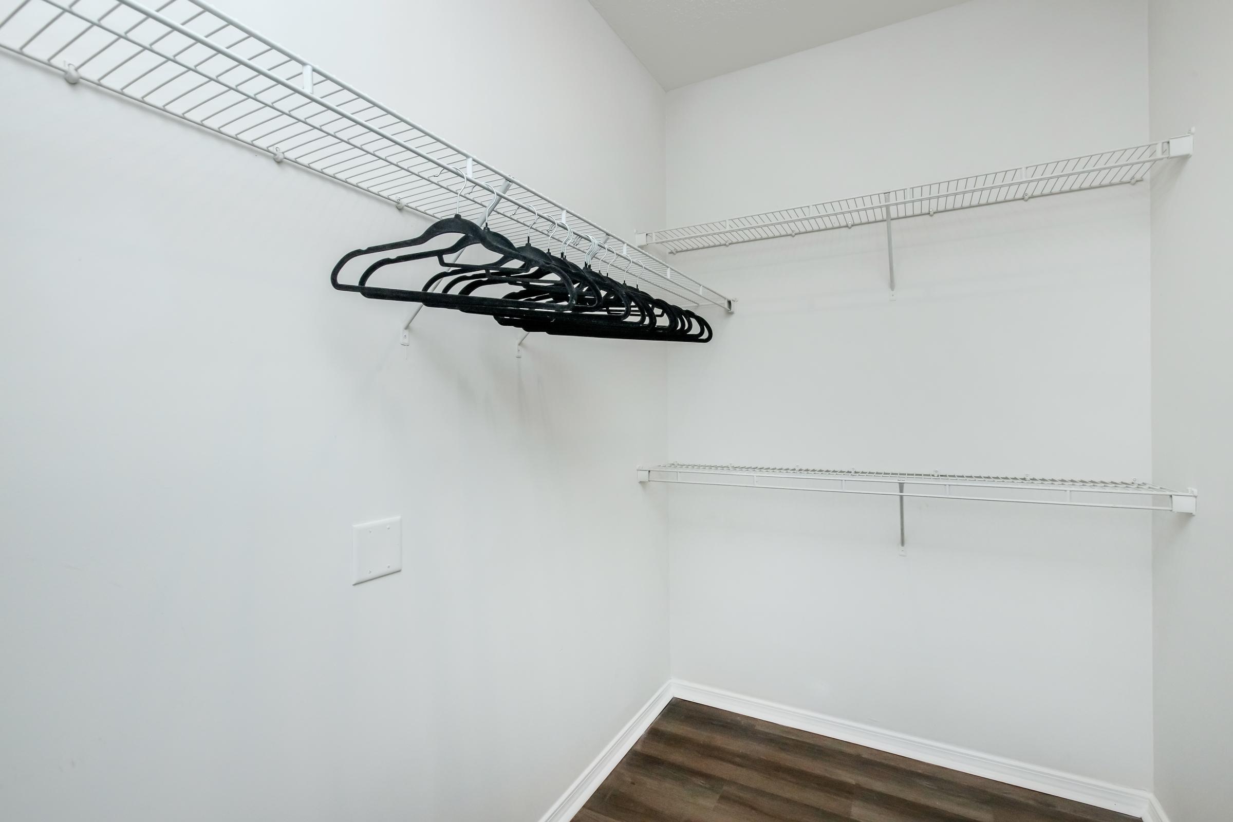Empty closet with white walls and a light wooden floor. There are metal wire shelves mounted on the walls, one on the left and one on the right. Black hangers are neatly hanging from the shelf on the left, while the shelf on the right is empty. The space is bright and uncluttered.