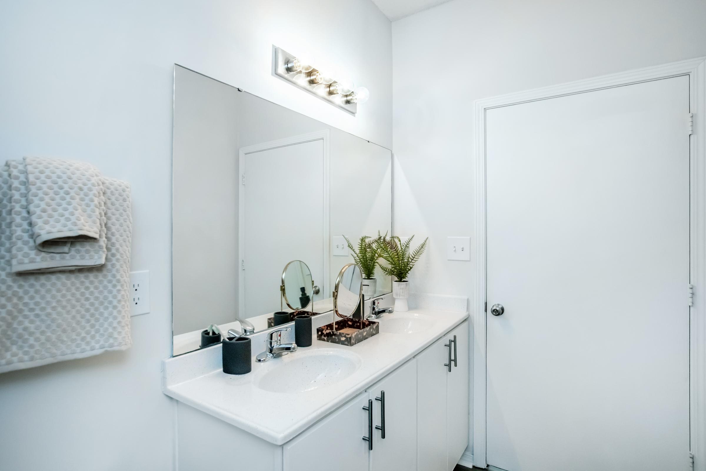 A modern bathroom featuring a white vanity with dual sinks, a large mirror, and a minimalist design. A towel hangs on the wall beside the sink, and a small potted plant adds a touch of greenery. The door is closed, and the overall color scheme is light and neutral.