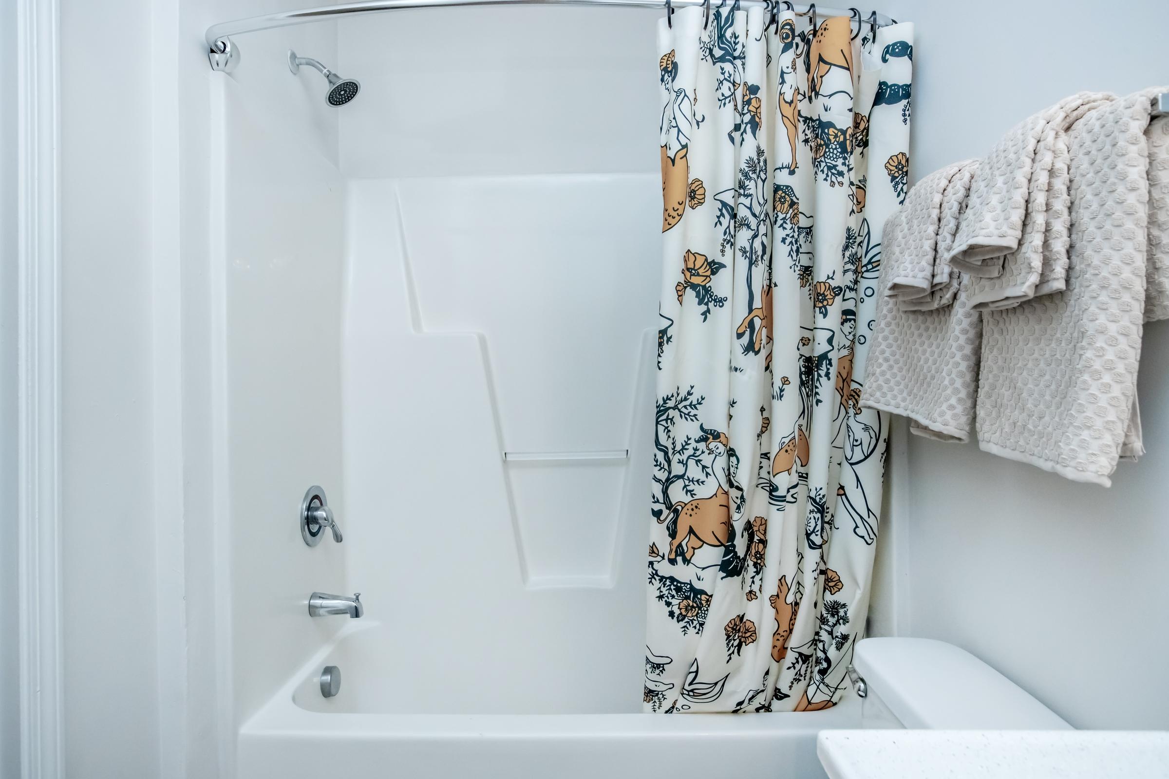 A clean bathroom featuring a bathtub with a shower. The shower curtain is decorated with animal illustrations and floral patterns. Two neatly folded beige towels hang on the wall nearby, adding a touch of warmth to the space. The overall decor is modern and minimalistic.