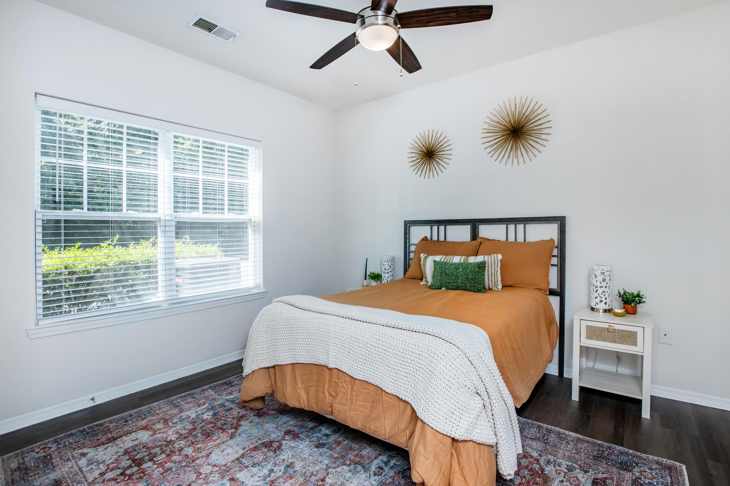 A cozy bedroom featuring a queen-sized bed with an orange bedspread and decorative pillows, a wooden headboard, and white nightstands with small plants. Two stylish wall decor pieces and a ceiling fan add a modern touch, while large windows provide natural light and a view of greenery outside.